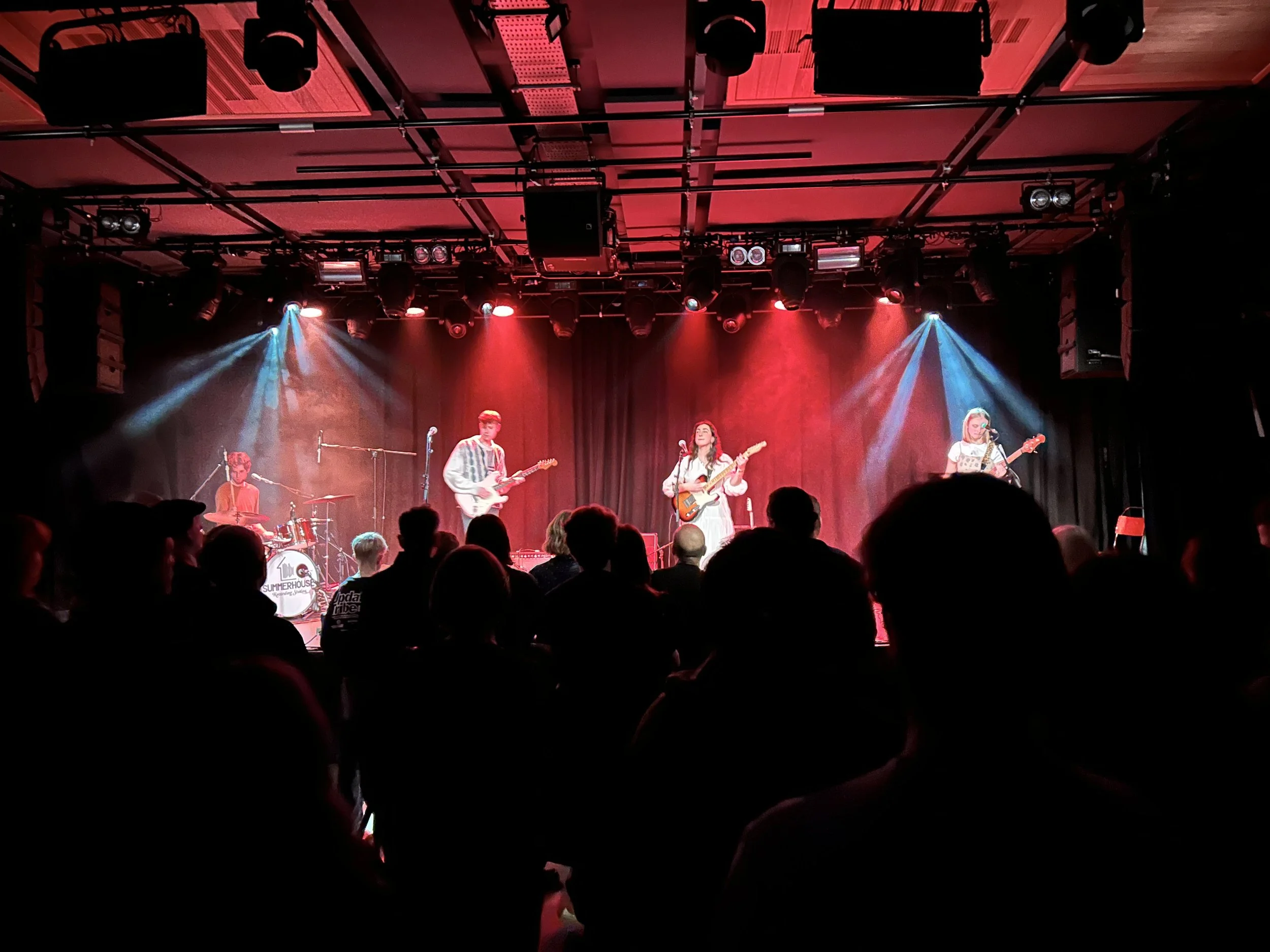 A band performs on stage with four members, including a drummer, two guitarists, and a vocalist, in front of an audience in a dimly lit venue with red and blue stage lighting.