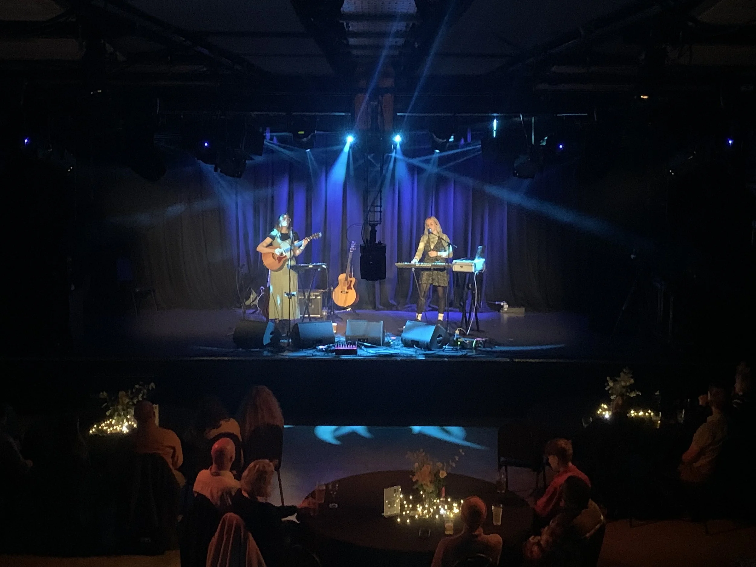 A live musical performance on stage with two female musicians, one playing guitar and singing, the other on a keyboard, illuminated by blue stage lights. Metronome Nottingham. Sunflower thieves.
