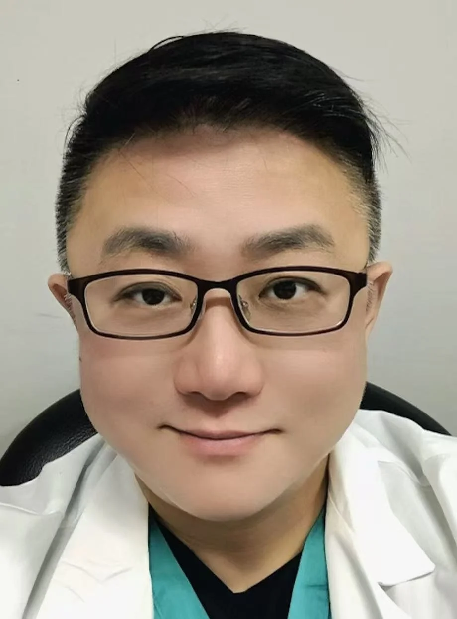 A young person with short black hair and glasses, smiling, wearing a white medical coat and teal scrubs, seated against a plain light-colored background.