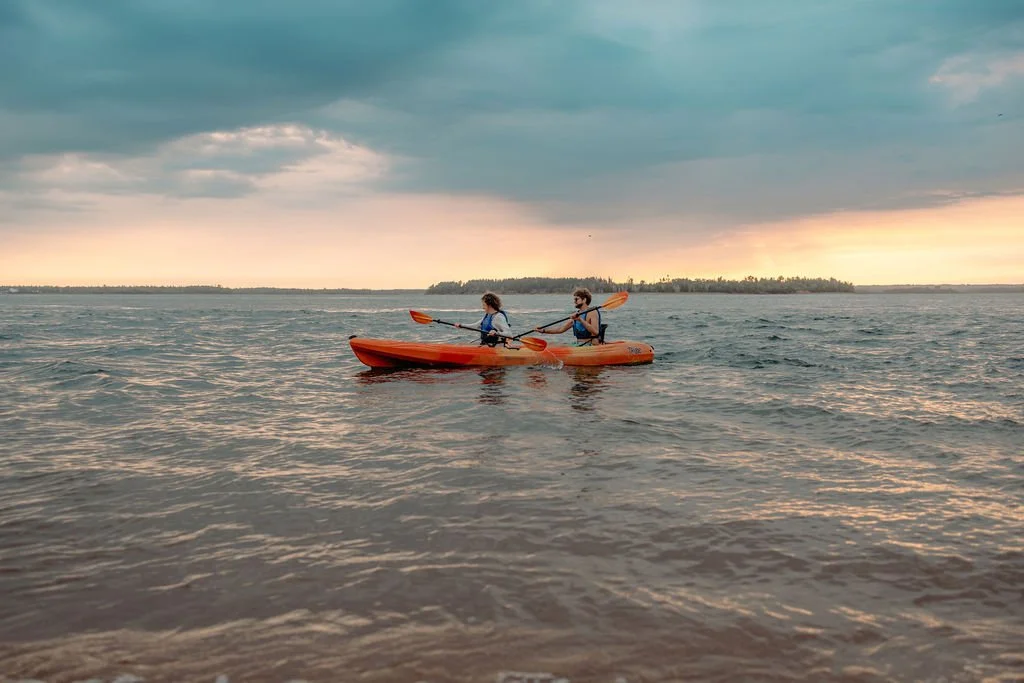 Two people kayaking in a lake during sunset, with overcast skies and distant shoreline.