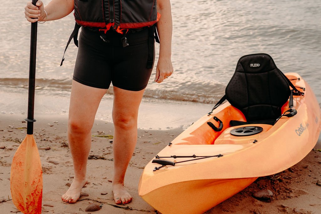 A person standing on a sandy beach holding a paddle next to an orange kayak with a black seat and a circular storage hatch.