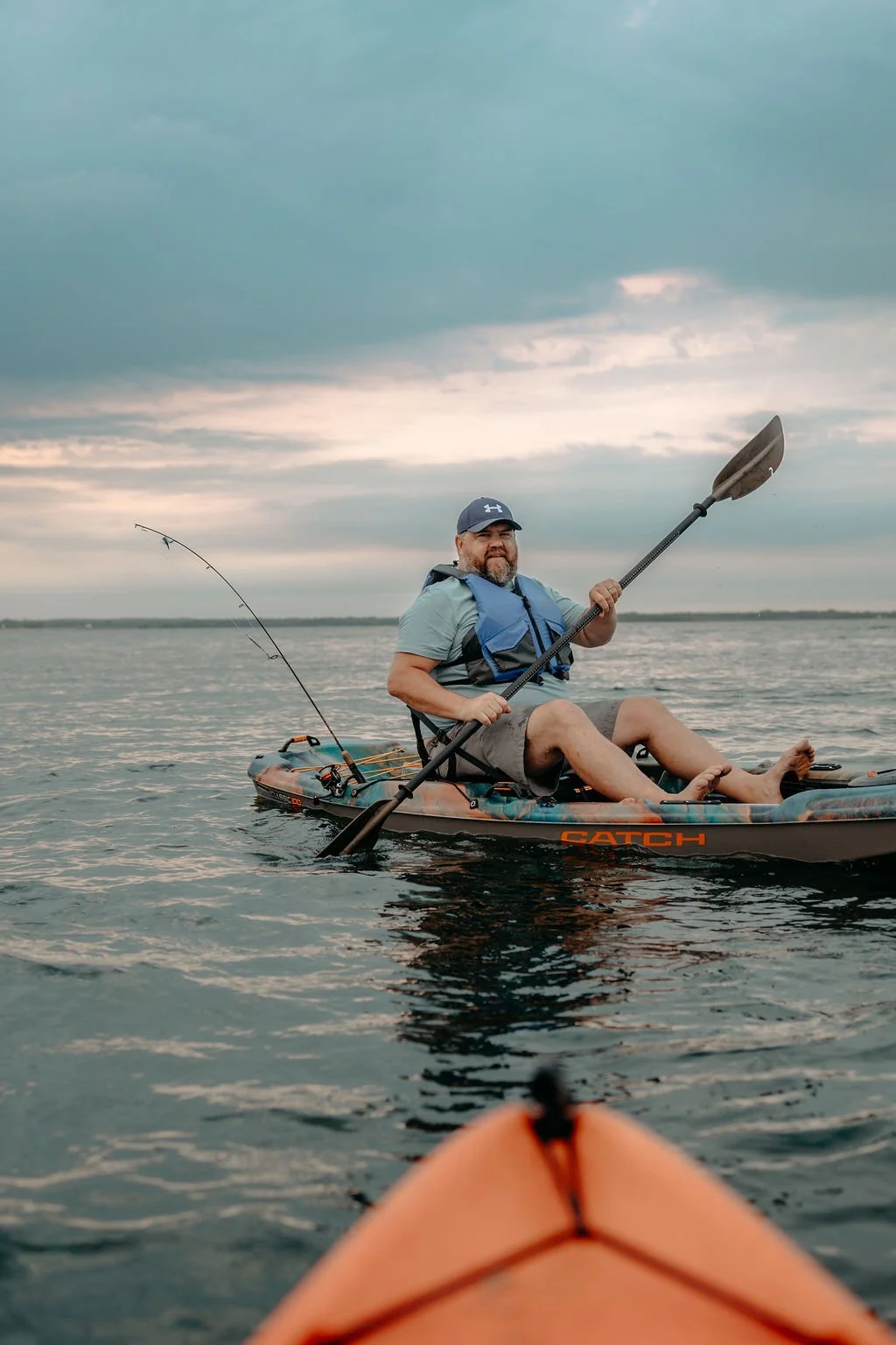 Man sitting in a kayak on a lake, holding a paddle, with fishing rods attached to the kayak, under a cloudy sky.
