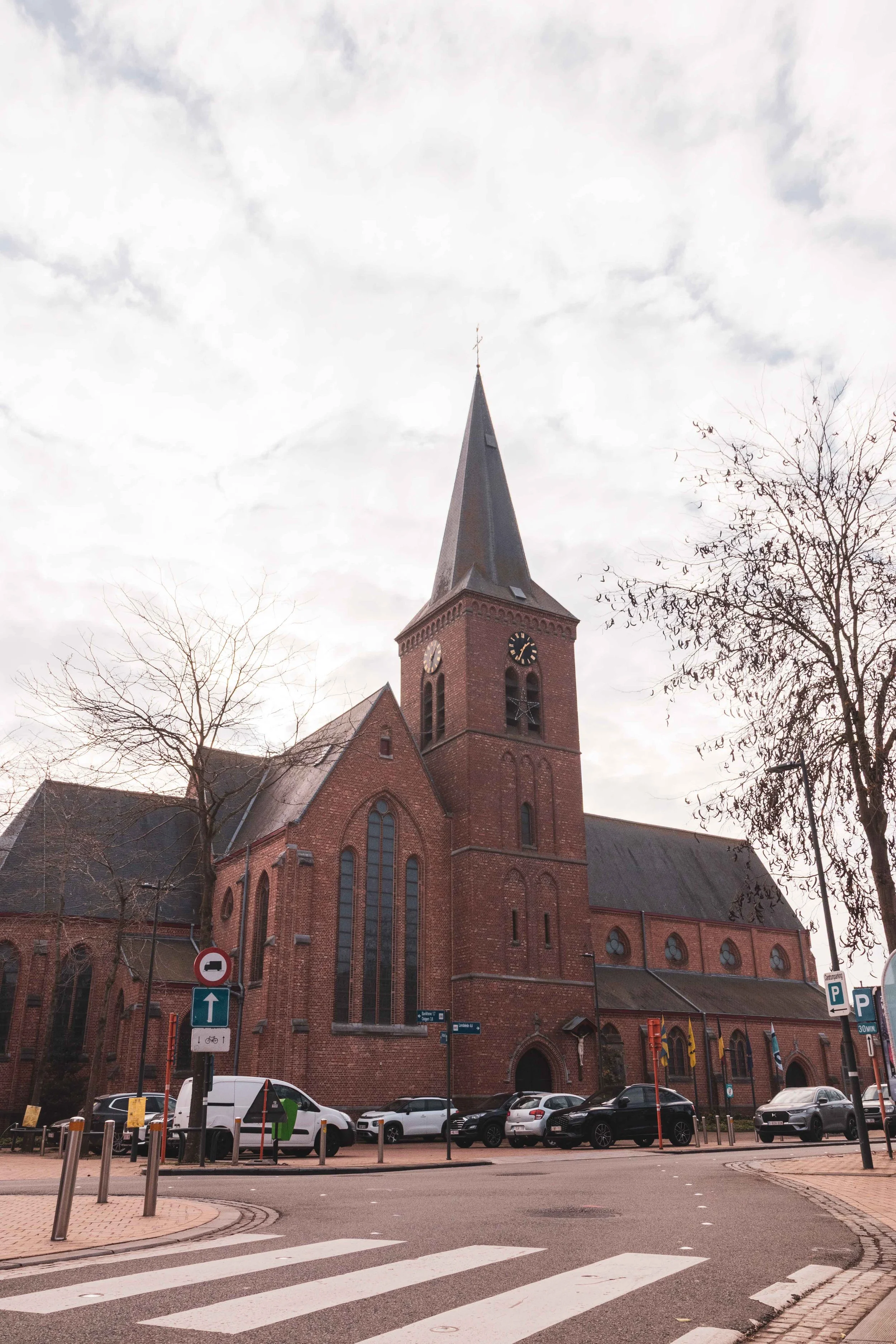 Een grote kerk gebouwd met rode bakstenen met een hoge spits, grote ramen en klokkentoren. Parkeerplaatsen en straatverkeer voor de kerk. Zonder publiek.