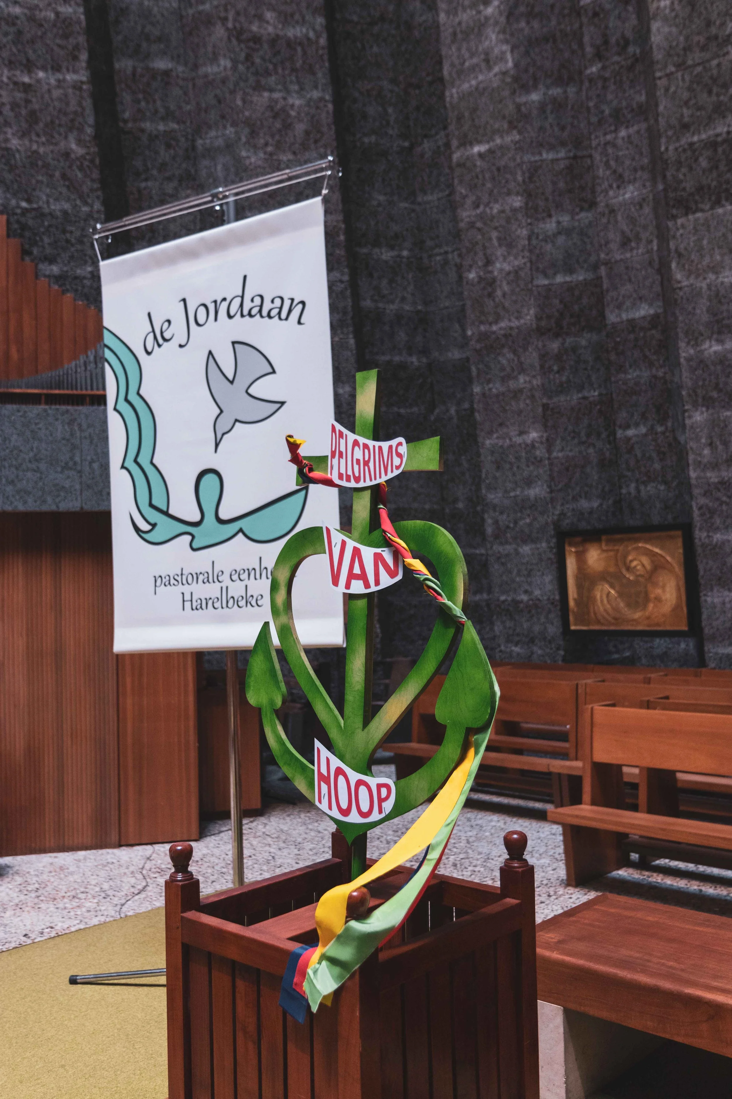 Een houten bloem met een bordje eraan met de woorden 'PELGIMS VAN HOP', in een kerk met houten banken en een beeldhouwwerk op de muur op de achtergrond.