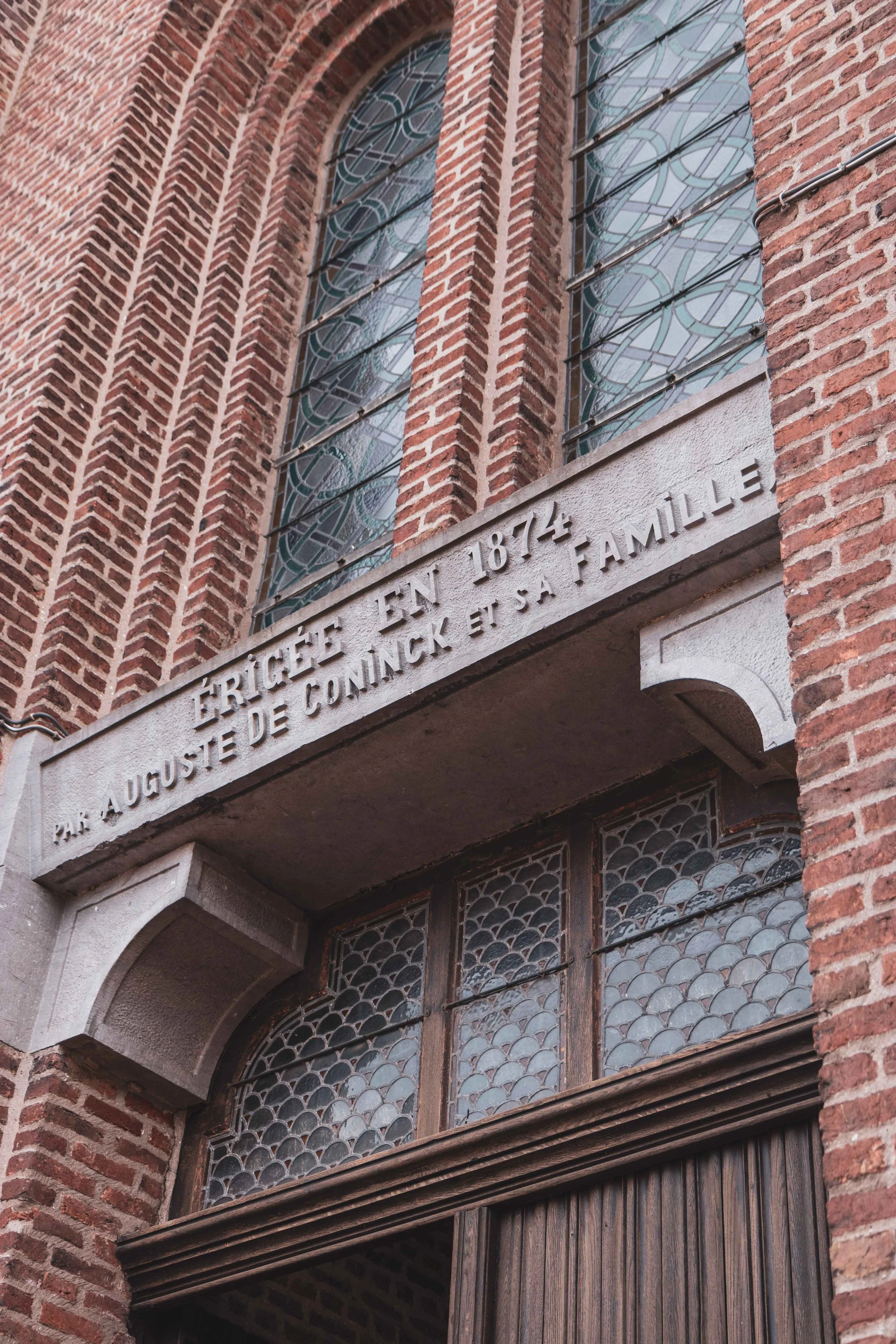 Gedenksteen op een bakstenen gebouw met sierglasramen, met inscriptie in het Frans over Auguste De Ginee en de Gennevite familie uit 1874.