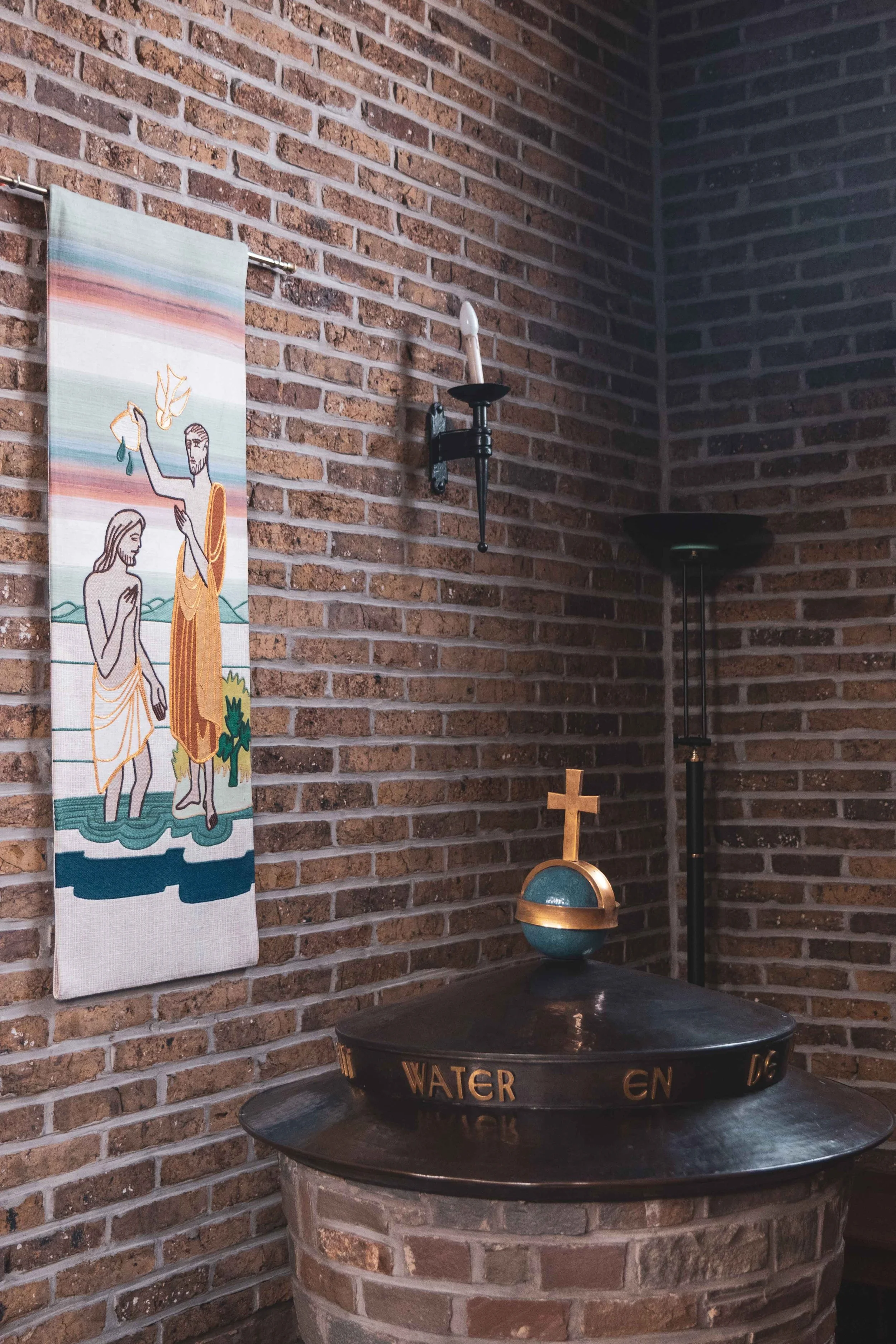 Een binnenruimte met een bakstenen muur, een hangende doek met een religieuze afbeelding, een muurhouder met een kaars, een staande lamp, en een ronde tafel met een decoratief ornament met een kruis en de tekst 'WATER en'.