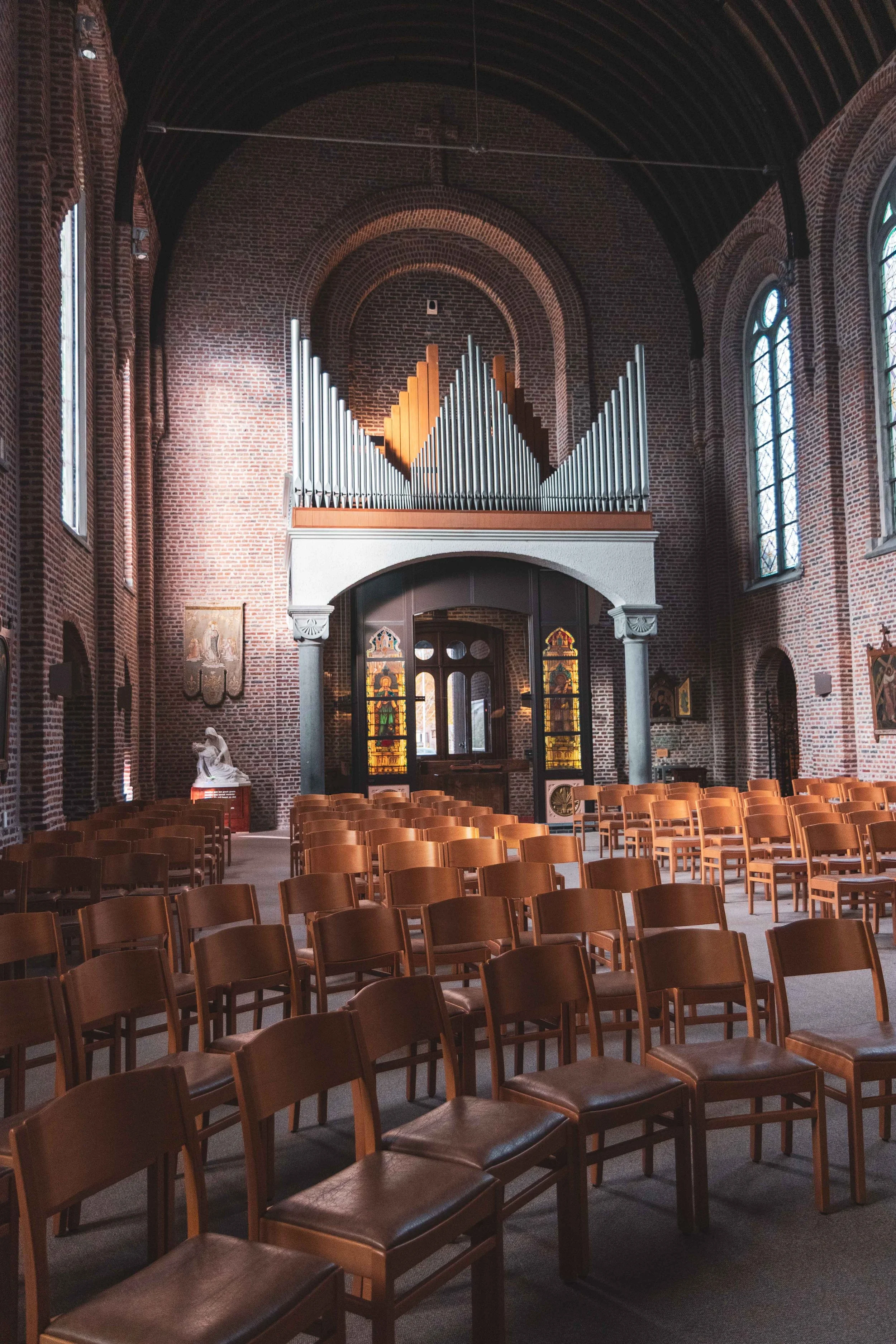 Interieur van een kerk met houten stoelen, een orgel boven de deur, en gebrandschilderde ramen.