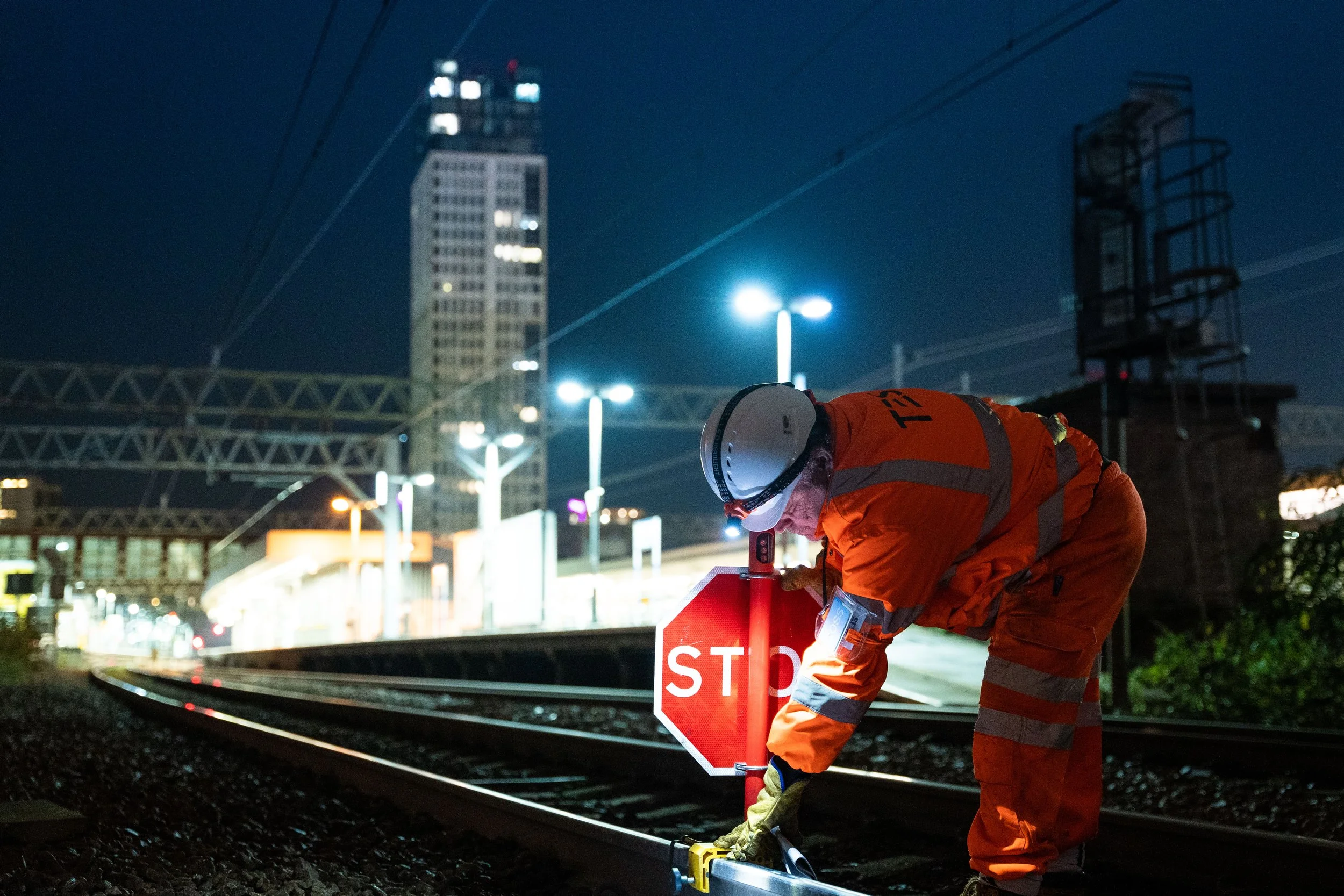 A worker in orange safety gear and a white helmet setting a small stop sign on railroad tracks at night, with tall buildings and city lights in the background.