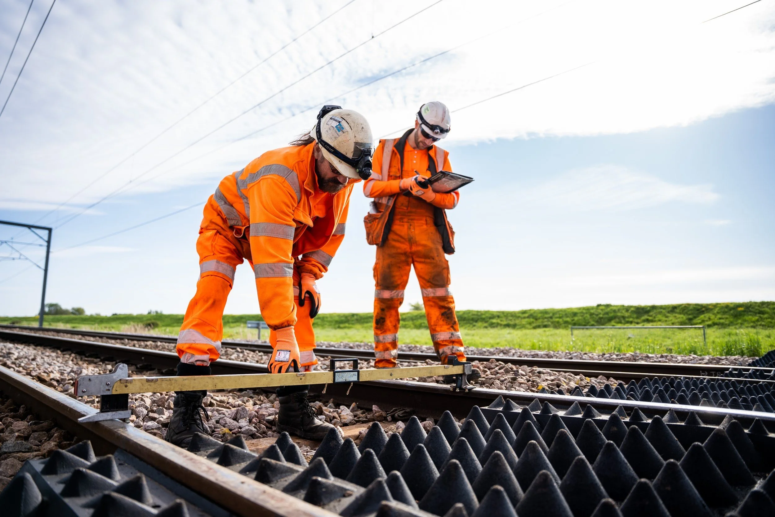 Two railway workers in orange safety uniforms and helmets inspecting railroad tracks outdoors on a clear day.