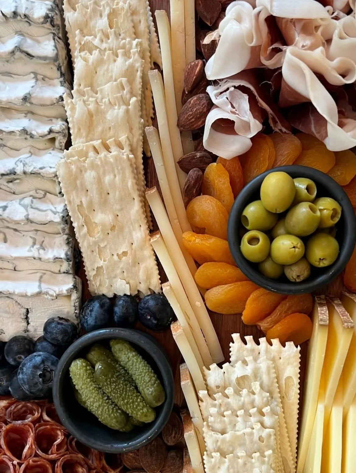 A snackboard filled with cheeses, olives, crackers, pickles, ham, dried apricots and blueberries.