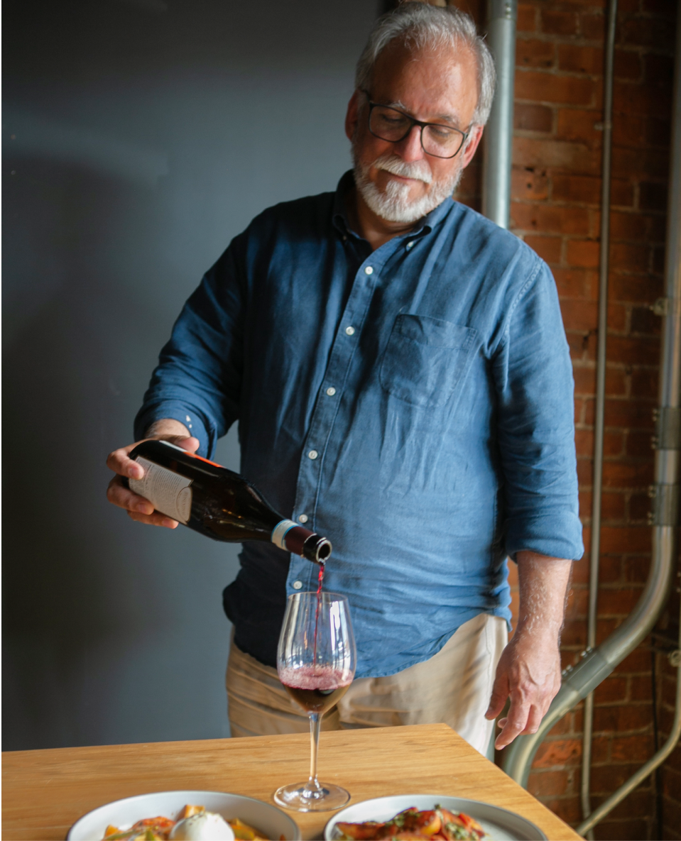 Older man with gray hair and beard pouring red wine into a glass at a table with plates of food, in a room with brick wall and pipes.
