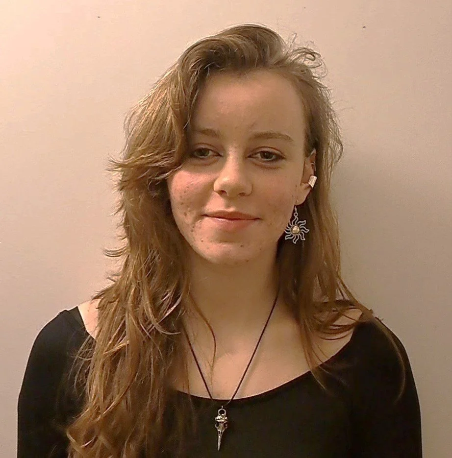 A young woman with wavy, light brown hair and light skin, wearing a black top, silver earrings resembling a sun and a moon, and a silver pendant necklace with a dagger, standing against a beige wall.