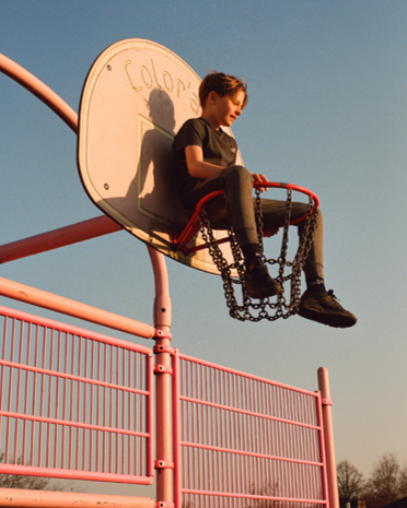 A girl sitting alone on a round, pink playground seat with chains, elevated above the ground, against a clear sky.