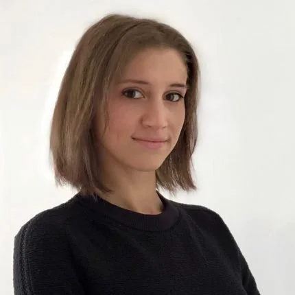 A young woman with shoulder-length brown hair wearing a black top, standing against a plain light background.