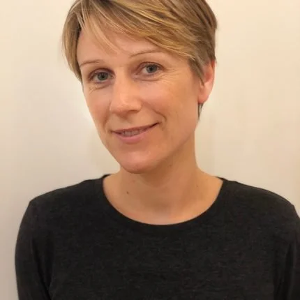 A woman with short blonde hair, wearing a black shirt, smiling at the camera against a plain light-colored background.