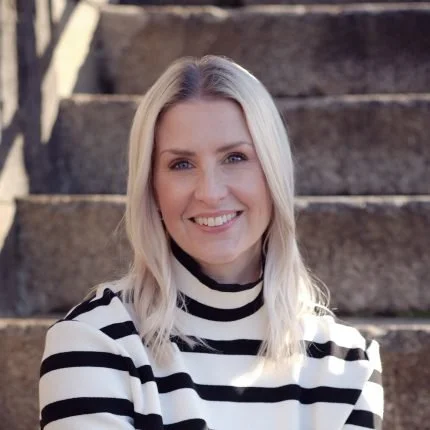 A woman with blonde hair smiling, wearing a black and white striped shirt, standing outdoors on stone stairs.