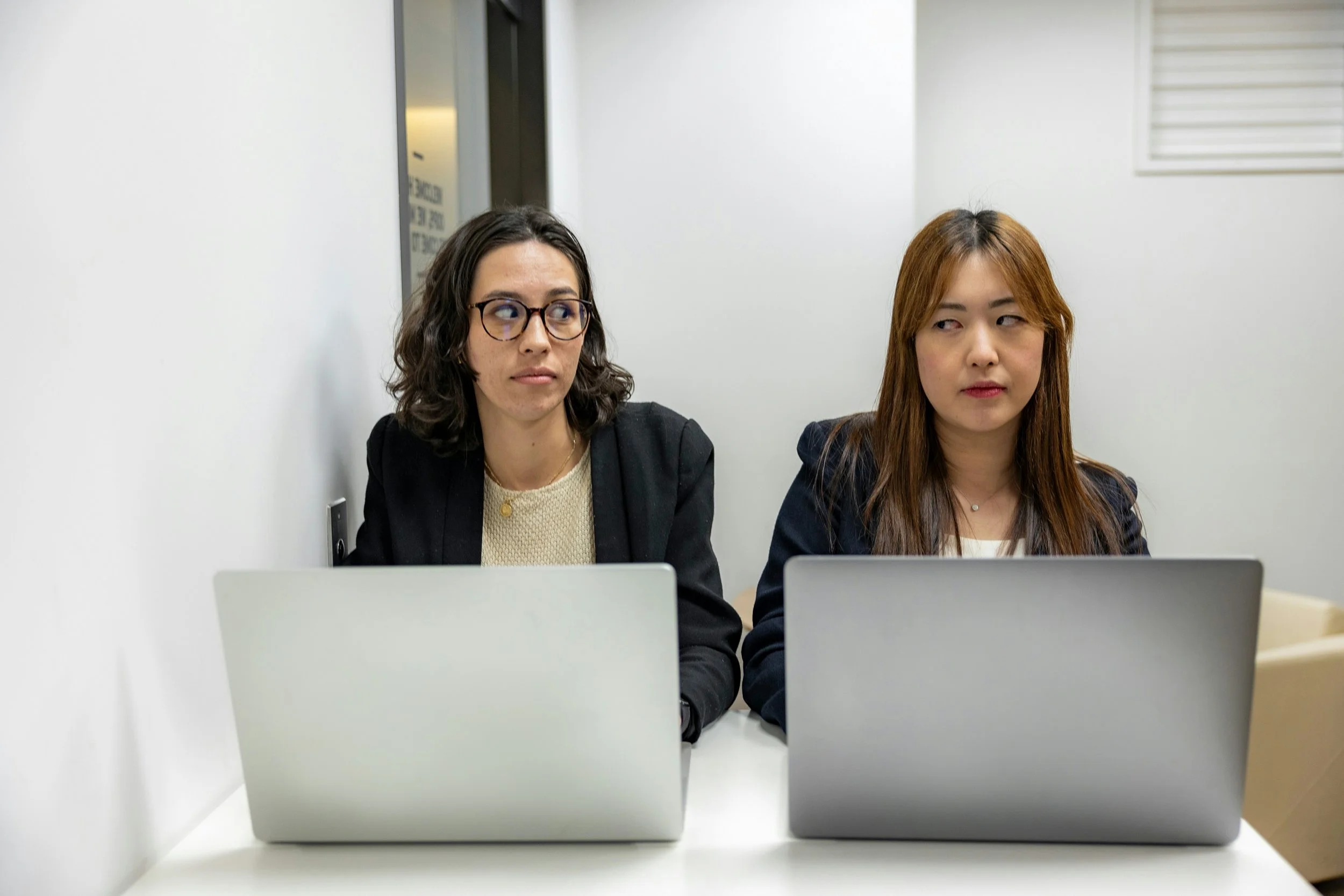 A white woman in her 30s with dark hair, glasses and in a black blazer is sitting next to an Asian woman in her 30s with long brown hair in a black blazer. Both have grey laptops in front of them and are looking ahead while side-glancing each other.