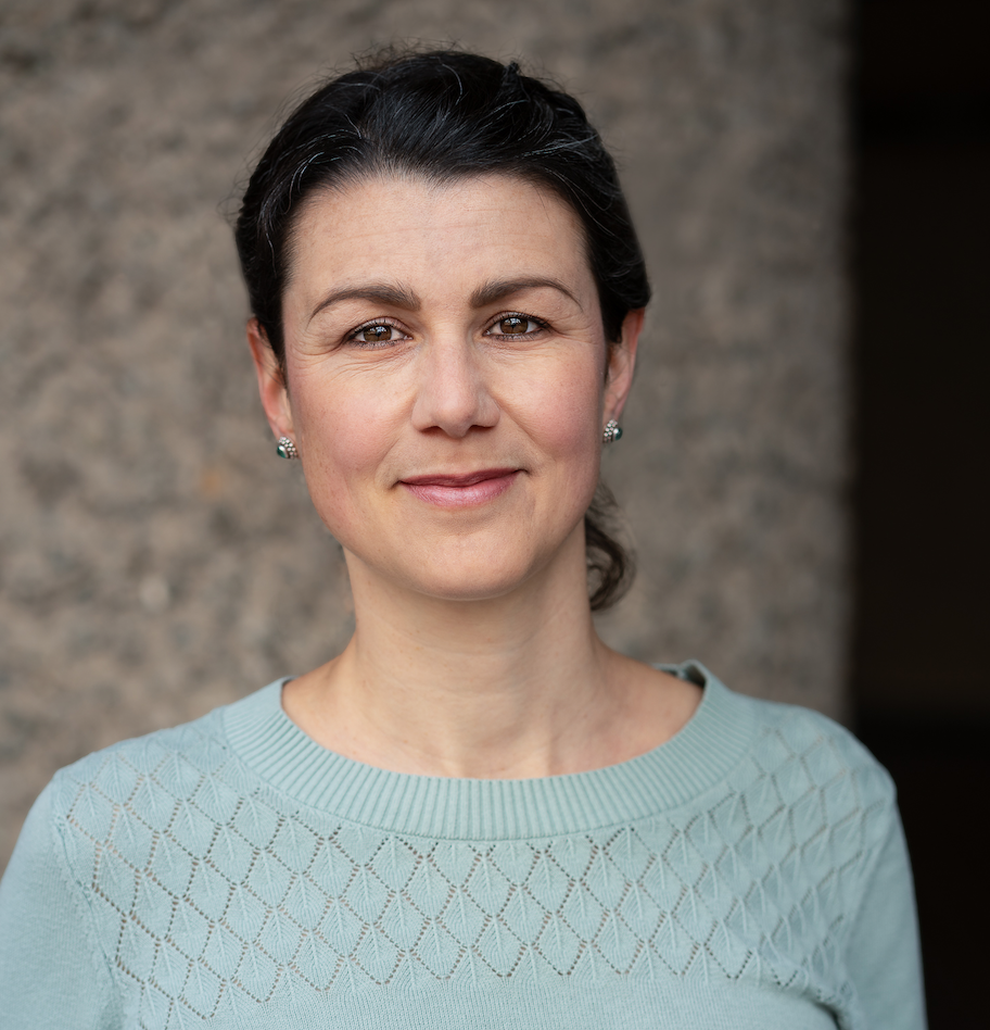 Headshot of Julia, a white woman in her 30s with dark hair braided into a bun is looking at the camera. She is standing in front of a dark grey background and wears a teal knitted top.