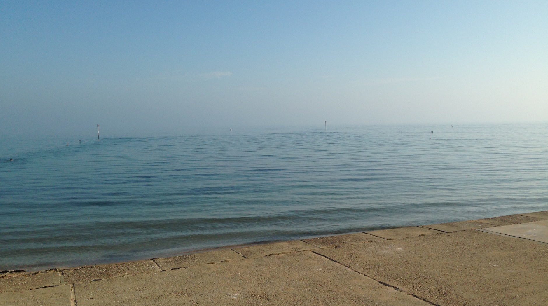 Photo of a sea pool in high tide, with the blue water submerging the poles that indicate the boundaries of the pool and partly submerging the brown-grey concrete slope leading into the water. The sky is a similar colour to the sea.