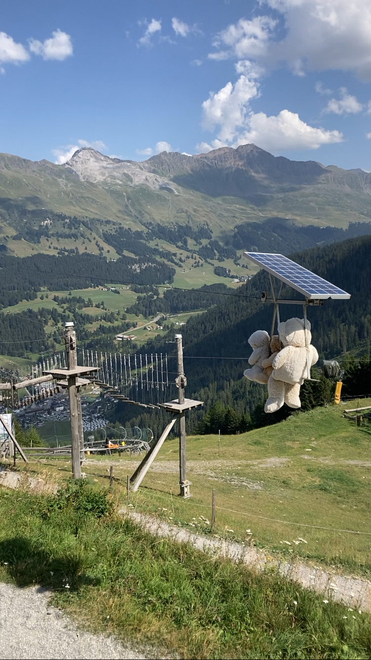 Photo of three teddy bears on a cable car powered by a tiny solar panel fixed above their heads overlooking a valley below flanked by cedar forests, green pastures and a rocky mountain range above in front of a blue sky.