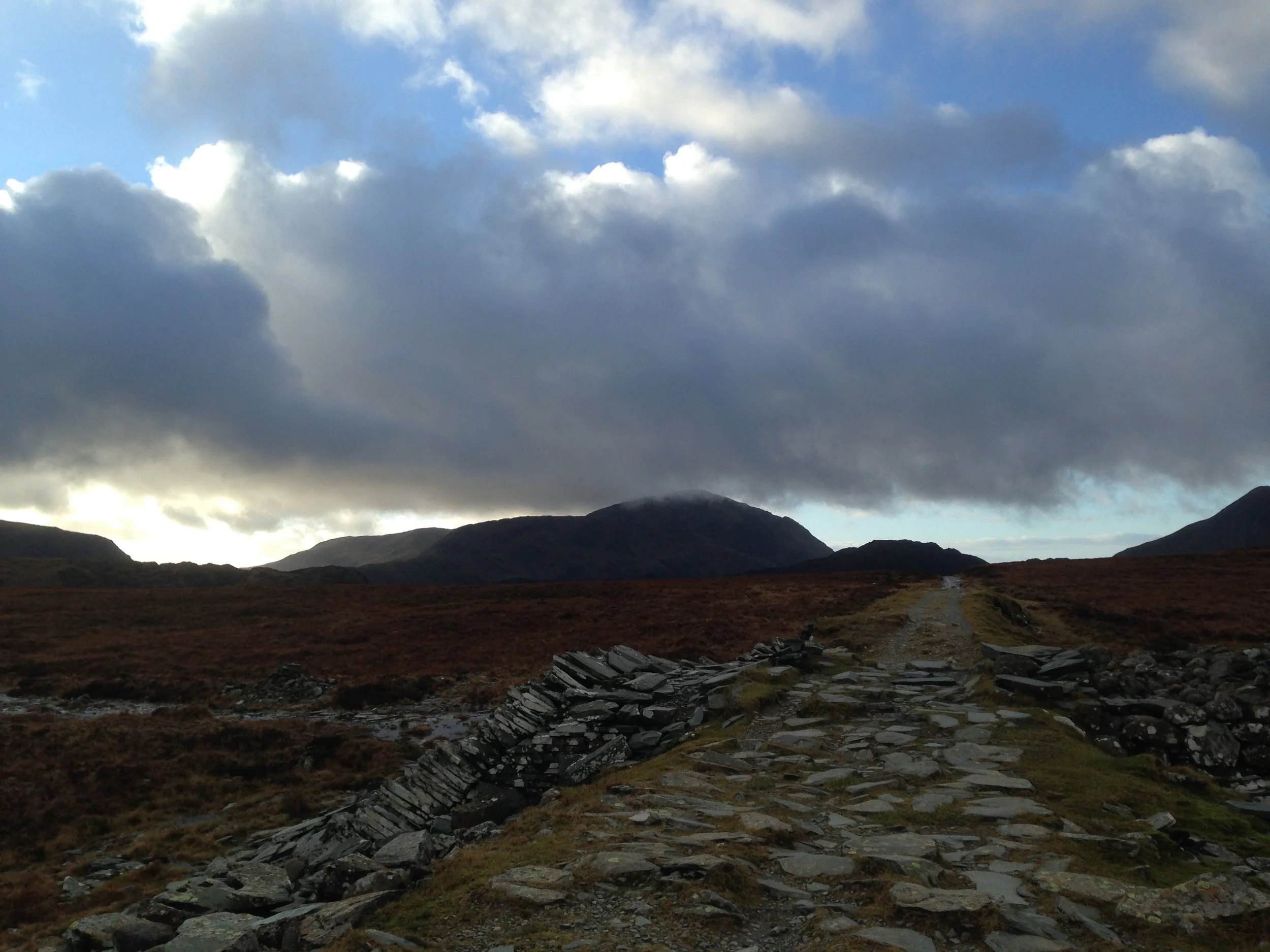 Photo of a footpath lined with flat rocks leading up a mountain with thick clouds overhead and some sunlight on the horizon, creating a dramatic mood