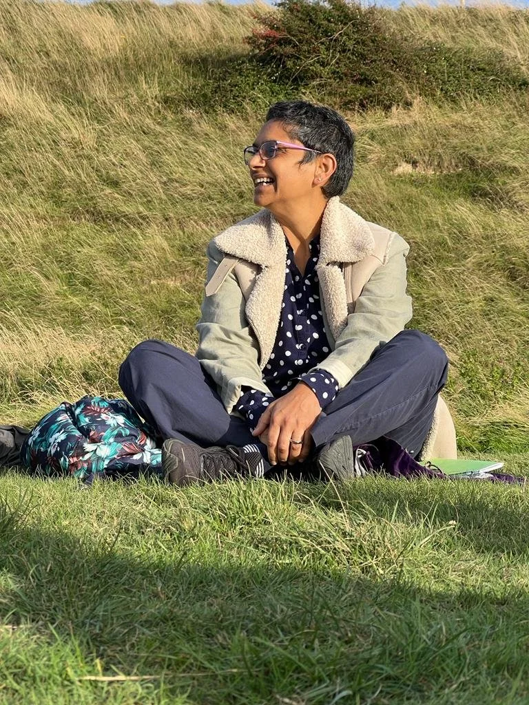 Razia is sitting on the grass in a beige coat and grey trousers with her legs crossed. She is laughing and looking over to the left side.
