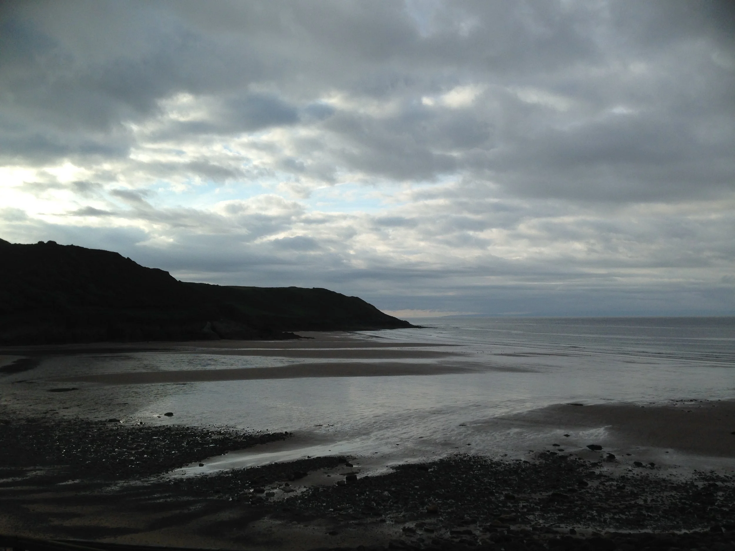 Photo of a beach at dusk, with grey clouds overhead, gentle waves on the ocean surface and dark cliffs framing the sea to the left side of the beach.