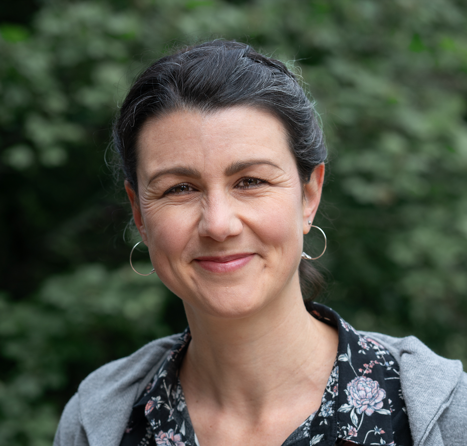 Headshot of Julia, a white woman in her 30s with dark hair braided into a bun smiling at the camera. She is standing in front of a green bush and wears a black blouse with floral paint and a grey cardigan on top.