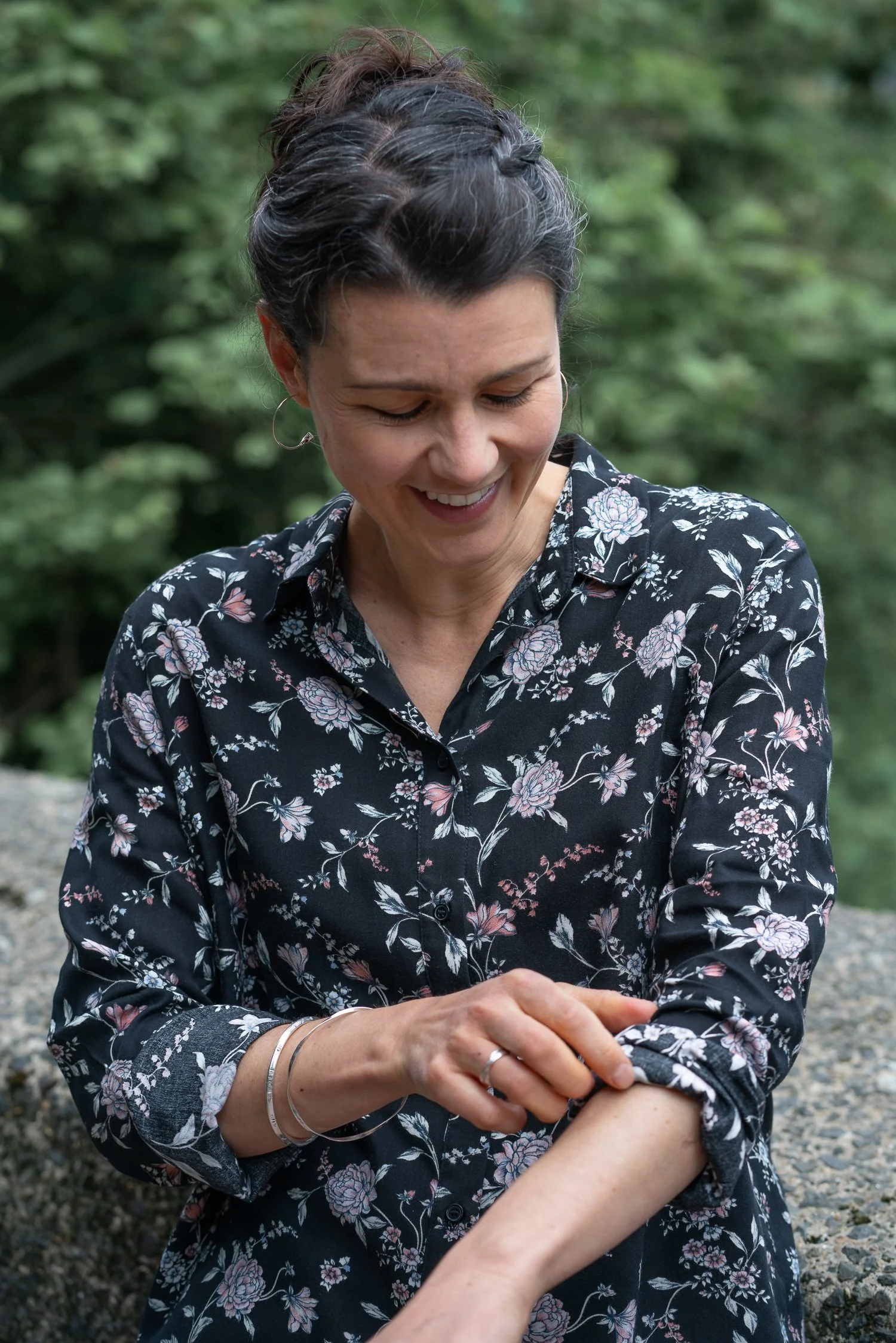Julia is smiling and looking down while rolling up the sleeves of her floral black blouse on her right arm.