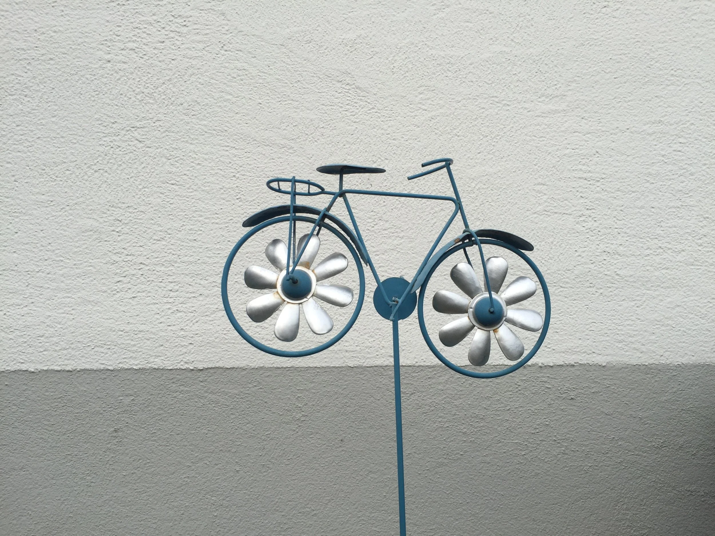 Photo of a decorative bicycle made of metal and painted in blue with silver flowers petals instead of spokes being held up on a thin blue stick in front of a grey wall.