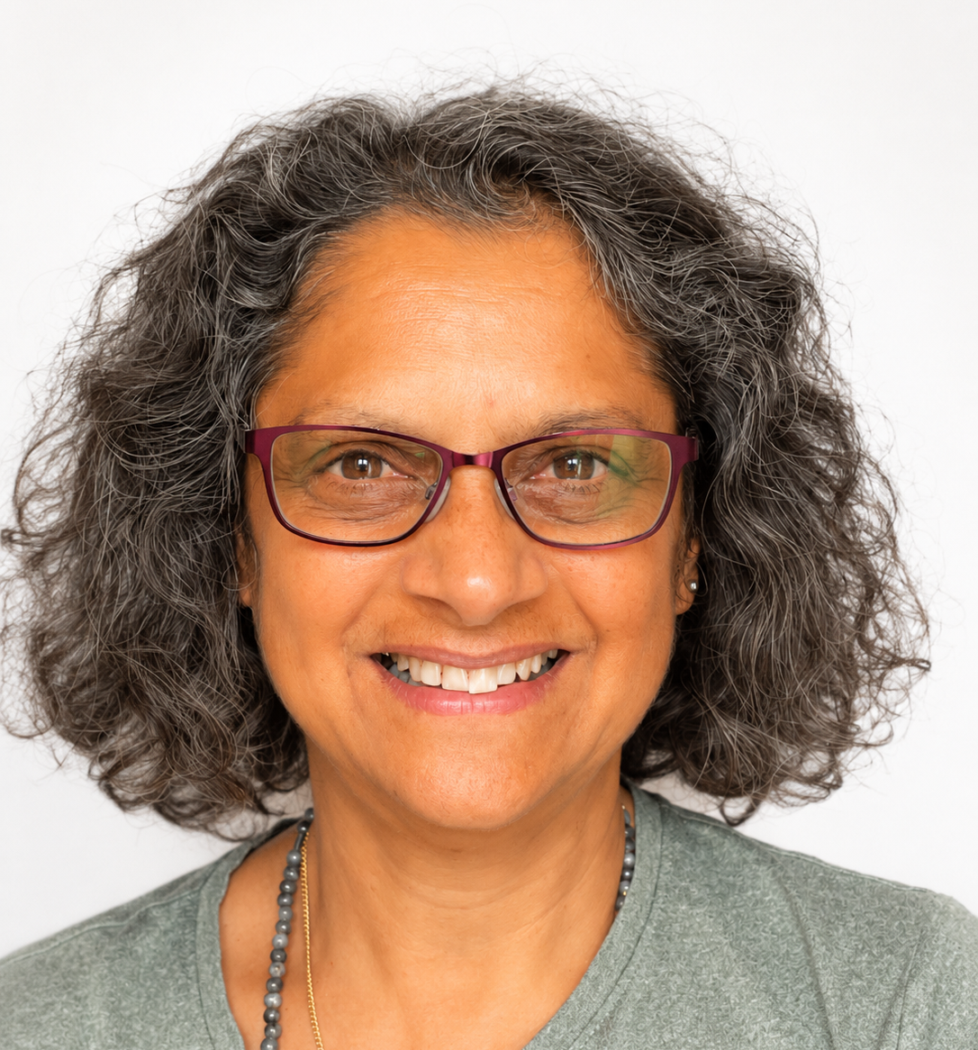 Headshot of Razia, a woman of Indian descent in her 60s with shoulder-length dark wavy hair and burgundy glasses in front of a white background. She is smiling at the camera and wears a grey top and matching necklace.