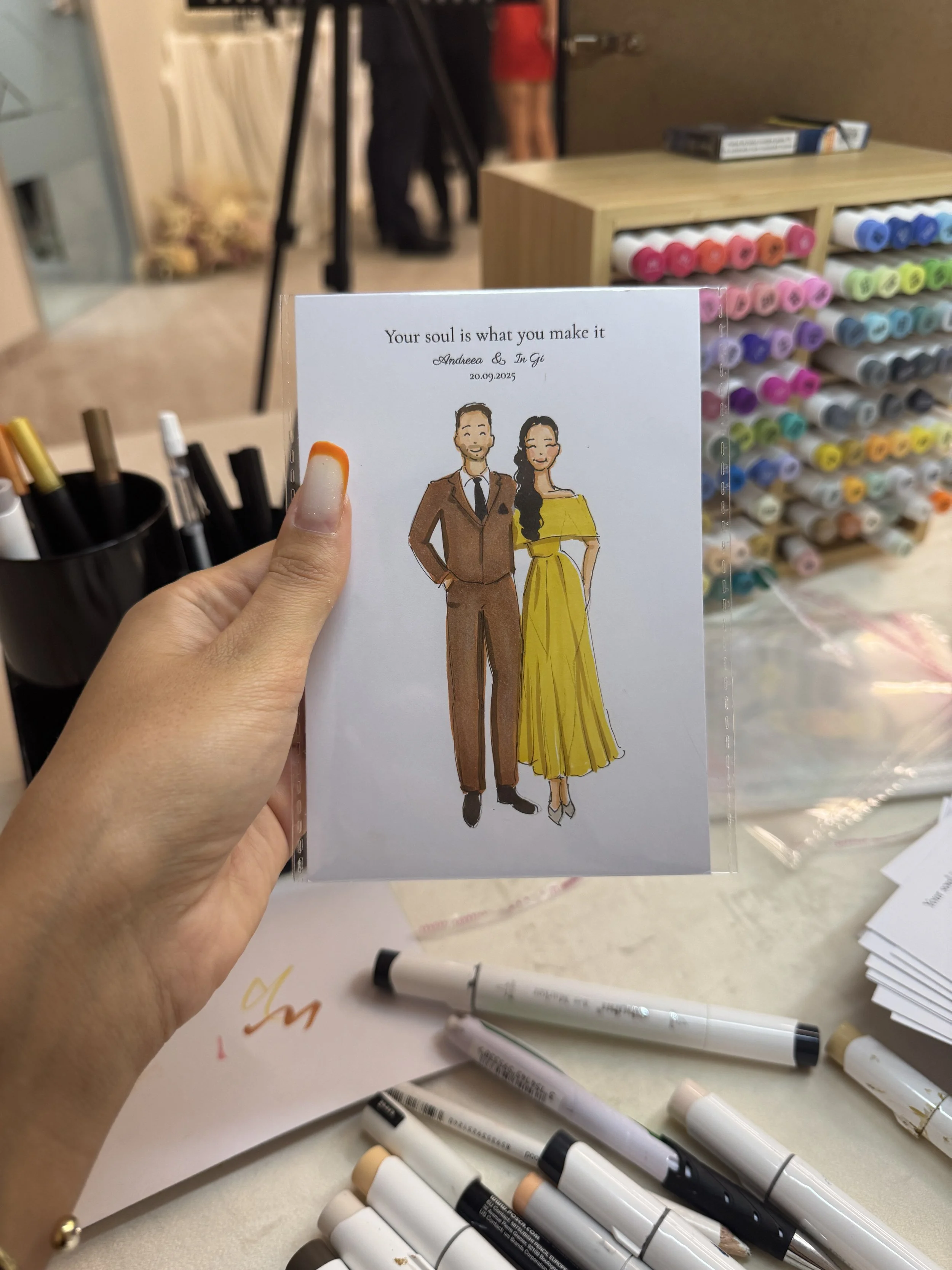 A person holding a wedding  illustration of a couple dressed in formal attire. The card has text that reads "Your soul is what you make it" and includes a date. In the background, there are colorful markers and a display with various colored pens or markers.