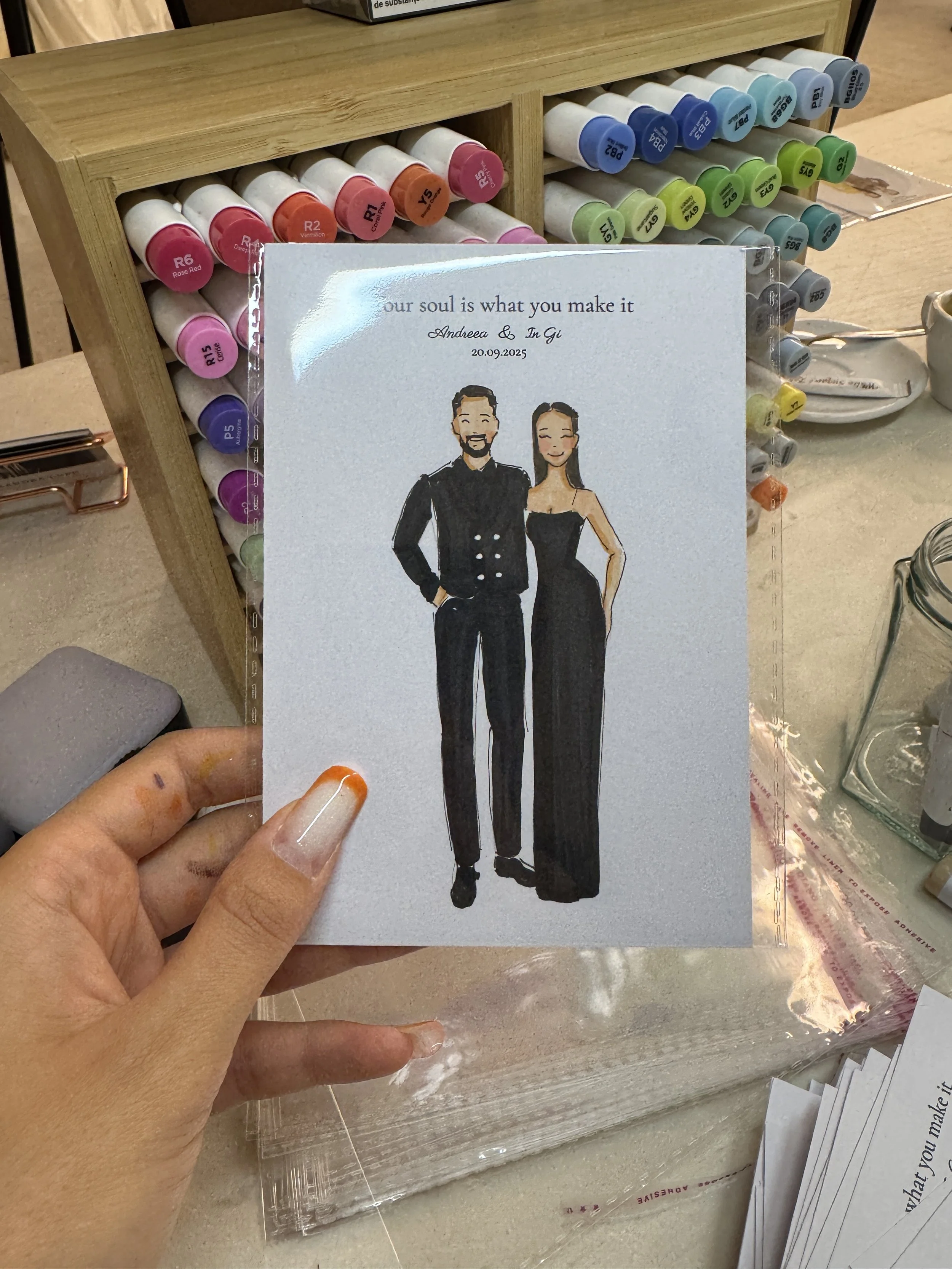A hand holding a card with an illustration of a couple in formal attire, set against a background of colorful markers and craft supplies.