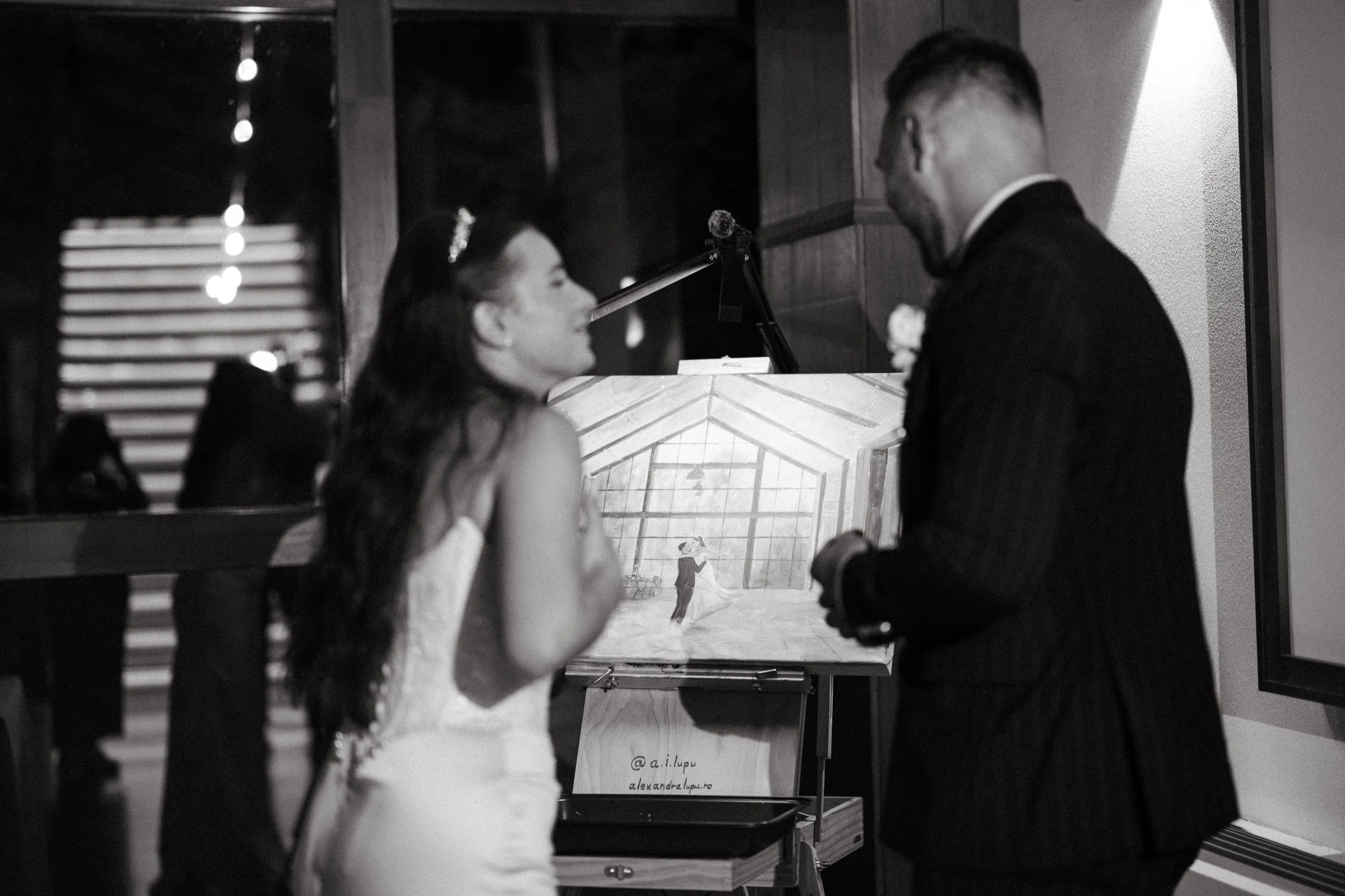 A bride and groom exchanging vows at their wedding, with the bride wearing a white dress and the groom in a dark suit, in a room with a wedding painting in the background.