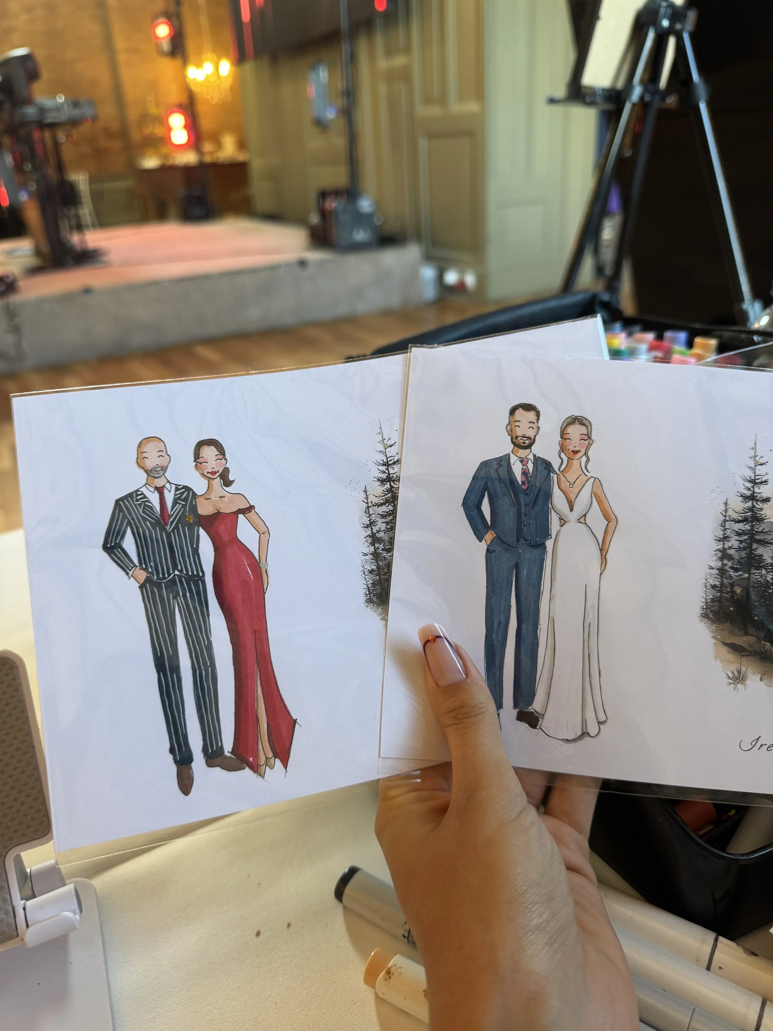 Two illustrated fashion models, a man and a woman, dressed in elegant formal attire, being held in a hand while sitting at a table in a dimly lit indoor setting.