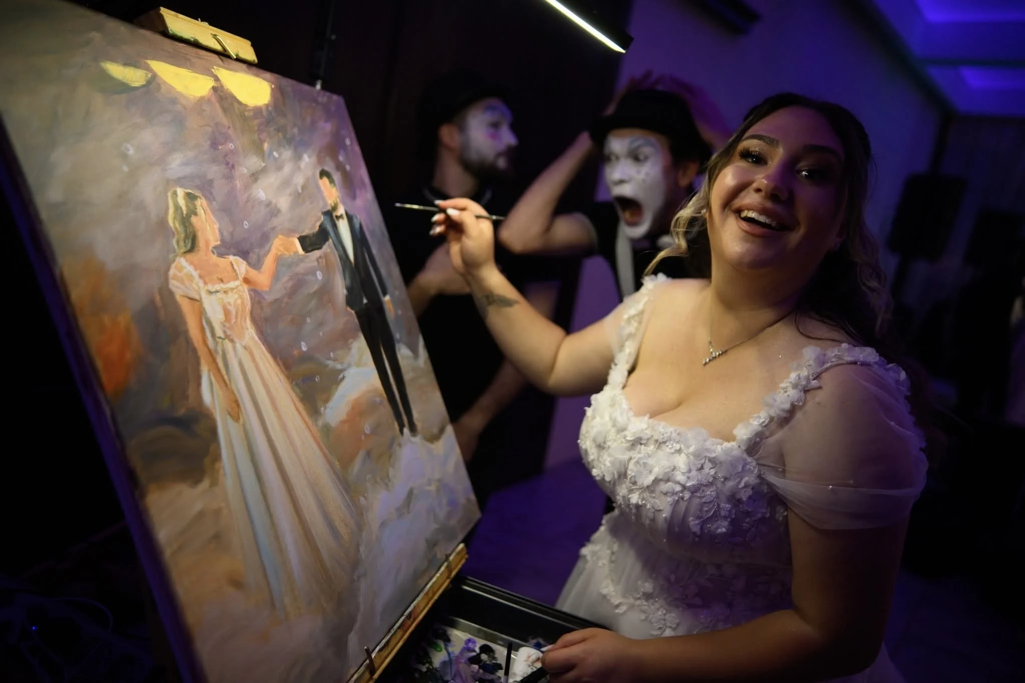 A bride painting a portrait of herself and the groom dancing, while two entertainers with painted faces perform in the background at an indoor event.