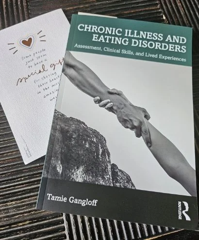 A book titled 'Chronic Illness and Eating Disorders' by Tamie Gangloff and a handwritten note with a small heart on white paper, both placed on a dark wooden surface.