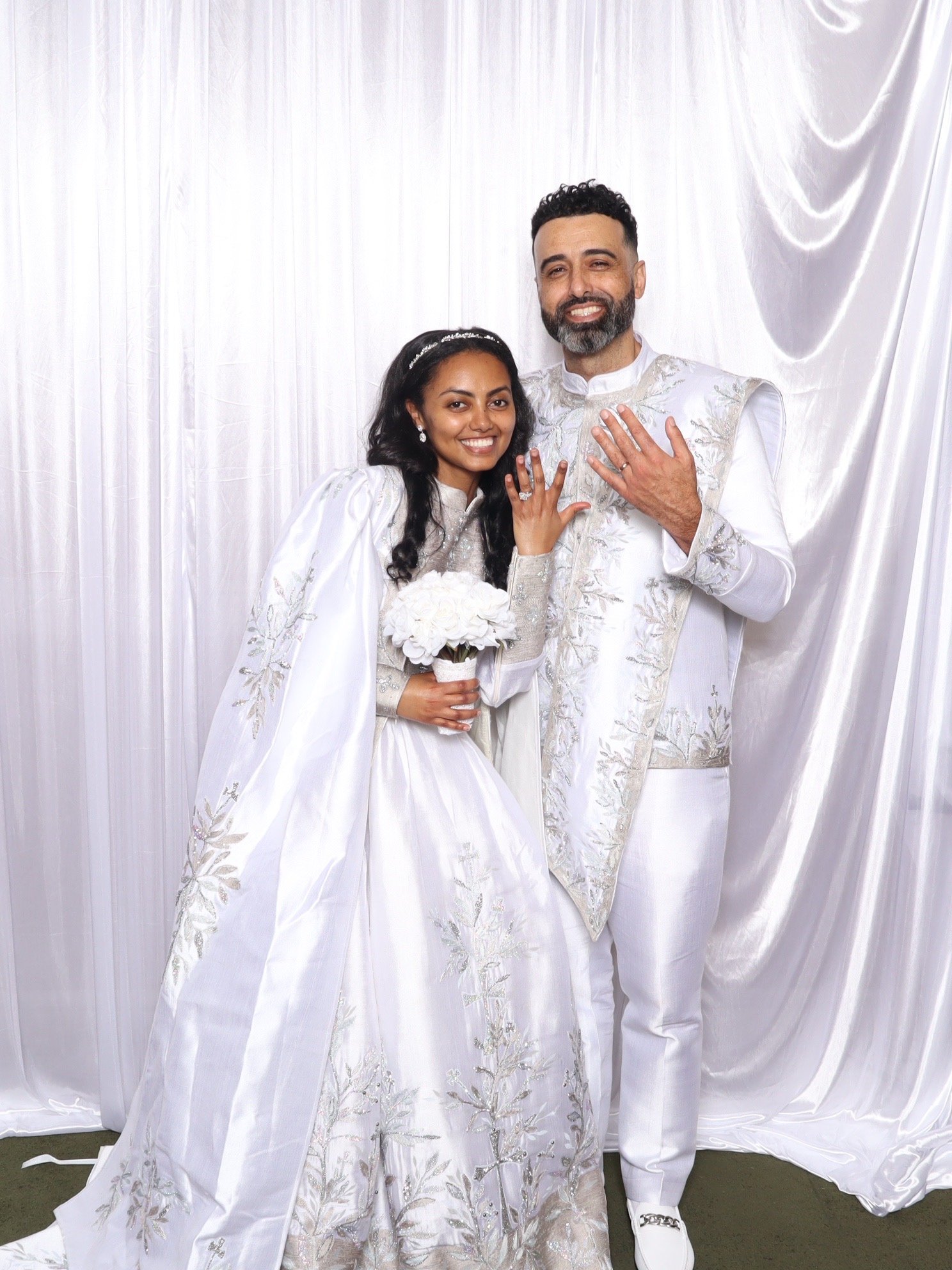A couple in white traditional attire, with the woman holding a bouquet, standing in front of a white draped background, showing their wedding rings and smiling.