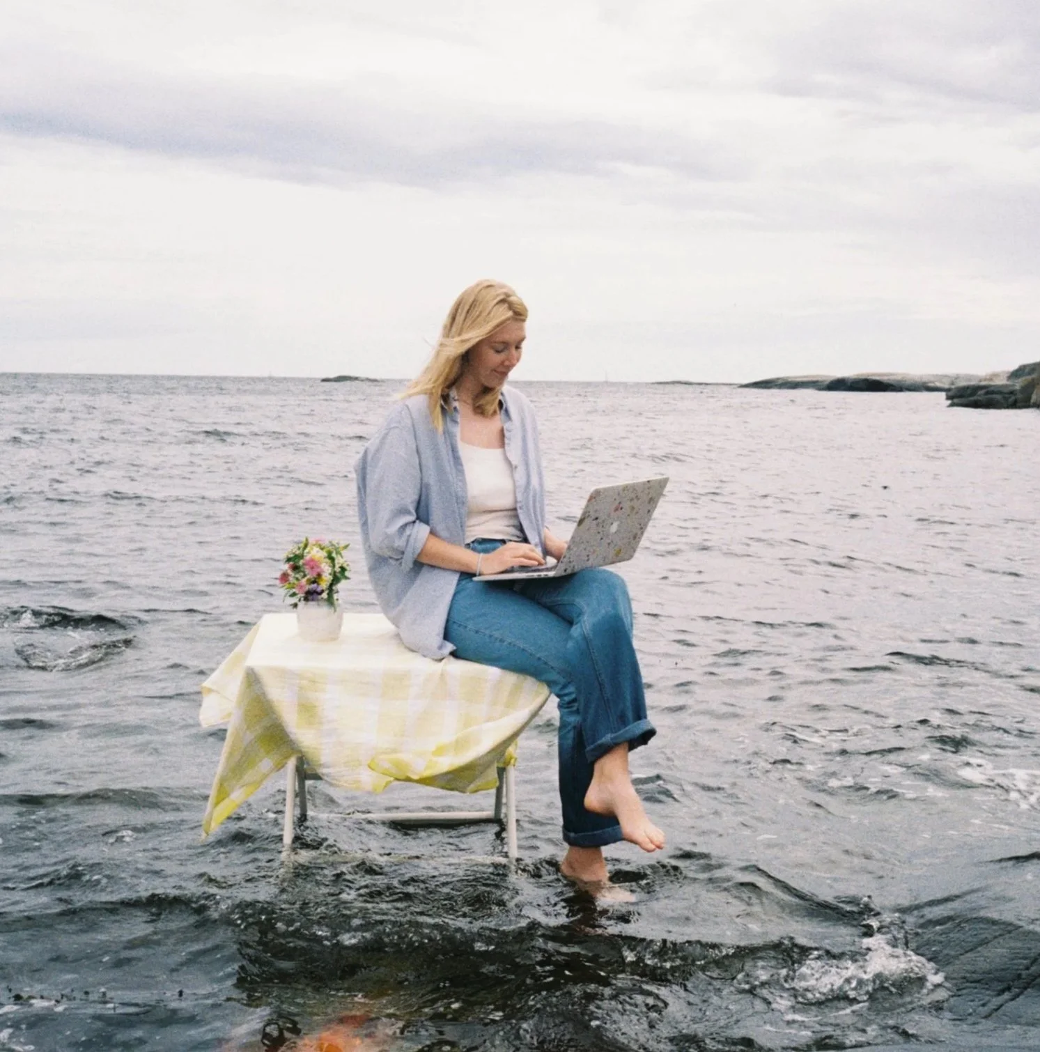 A woman sitting on a small table placed in the shallow water, with a yellow checkered tablecloth, working on a laptop, with her feet dipped in water by the ocean on a cloudy day, and a small flower arrangement beside her.