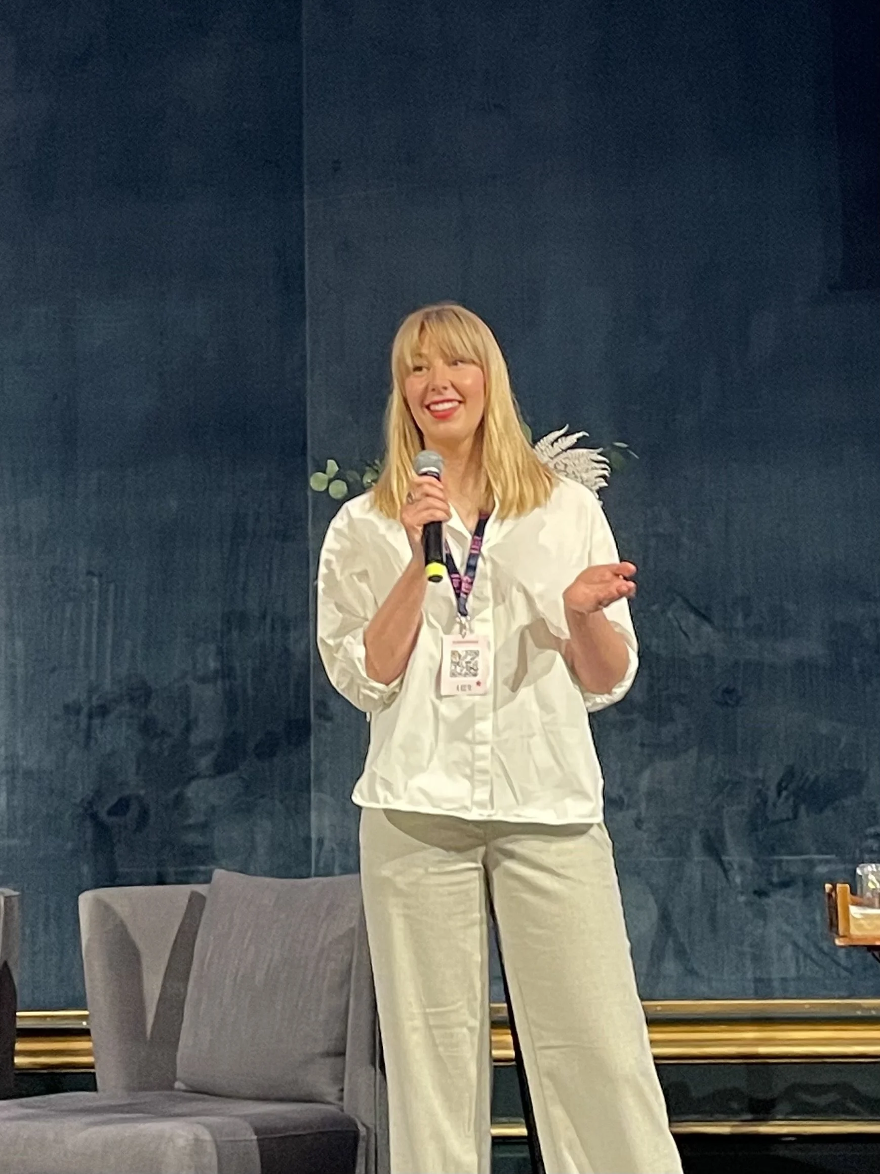 A woman with blonde hair wearing a white shirt and beige pants standing on stage, holding a microphone, smiling, and speaking at an event with a blue background and chairs nearby.