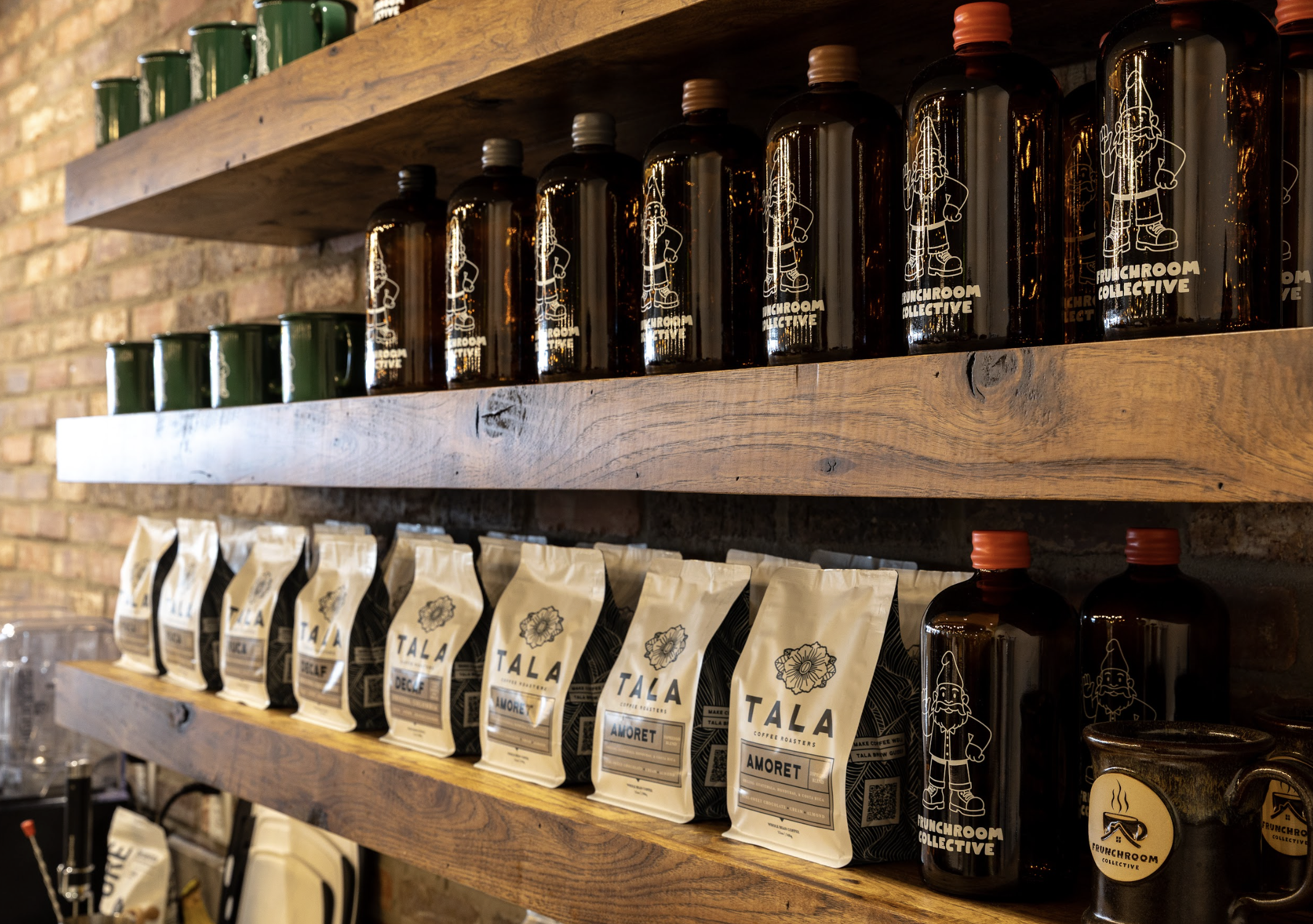 Wooden shelves displaying bottles with a logo of a gnome and coffee-related labels, along with green cans and beige coffee packaging, set against a brick wall background.