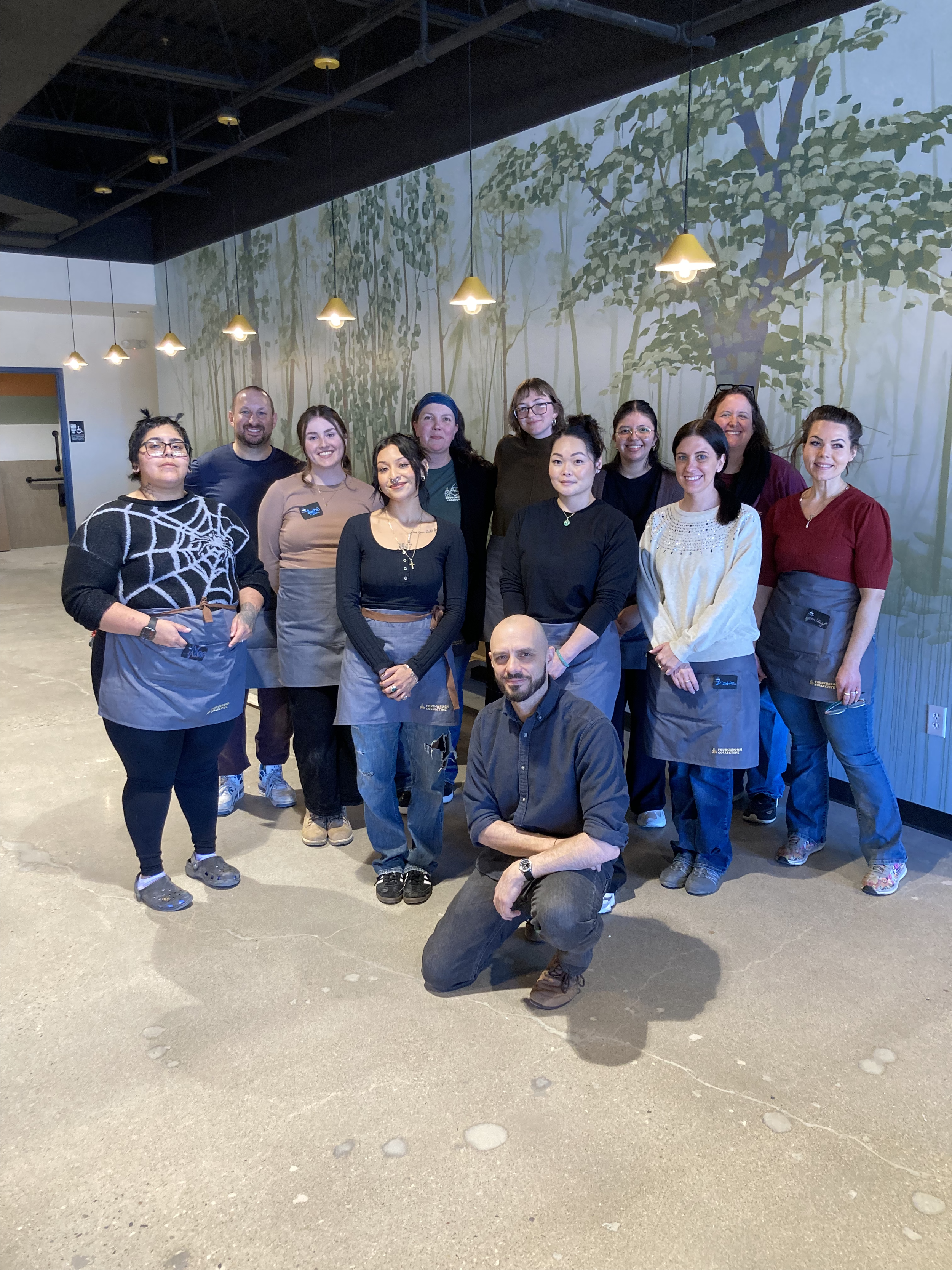 Group of people posing in front of a painted nature-themed mural, some wearing aprons, indicating they might be staff or attendees at a workshop or event.