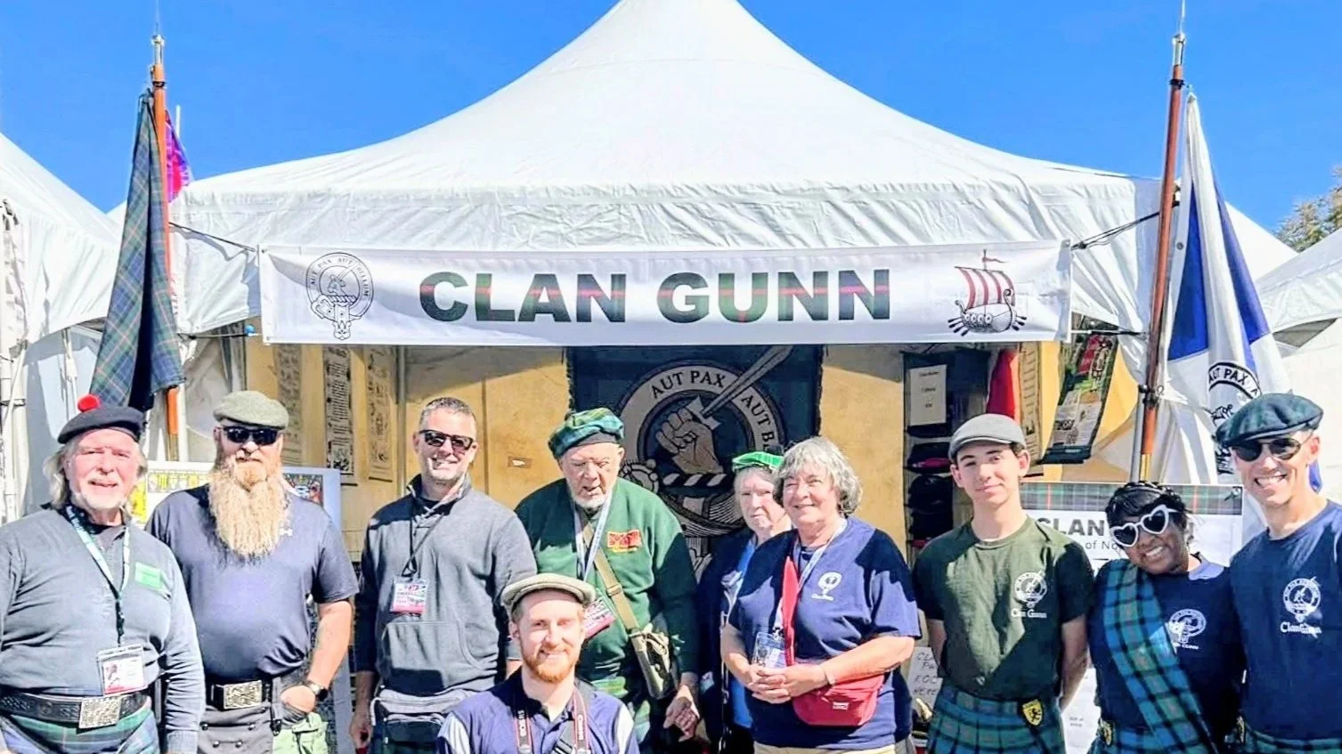 A group of people standing in front of a Clan Gunn booth at an outdoor event, with banners and flags, under a blue sky.