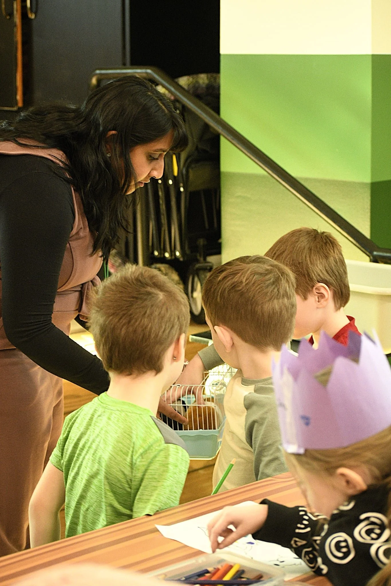 A woman with dark hair leaning over a group of children sitting at a table, engaged in an activity. The children have light hair and are focused on the task. The scene appears to be in a classroom or indoor learning environment, with stacked chairs and a green and cream wall in the background.