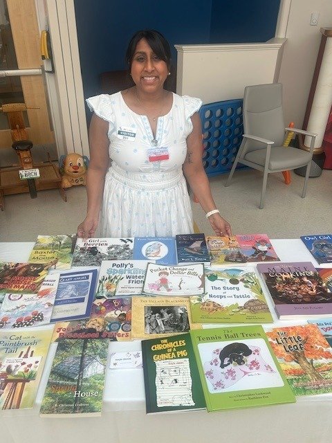 Woman smiling behind a table filled with children's books at a book sale or event.