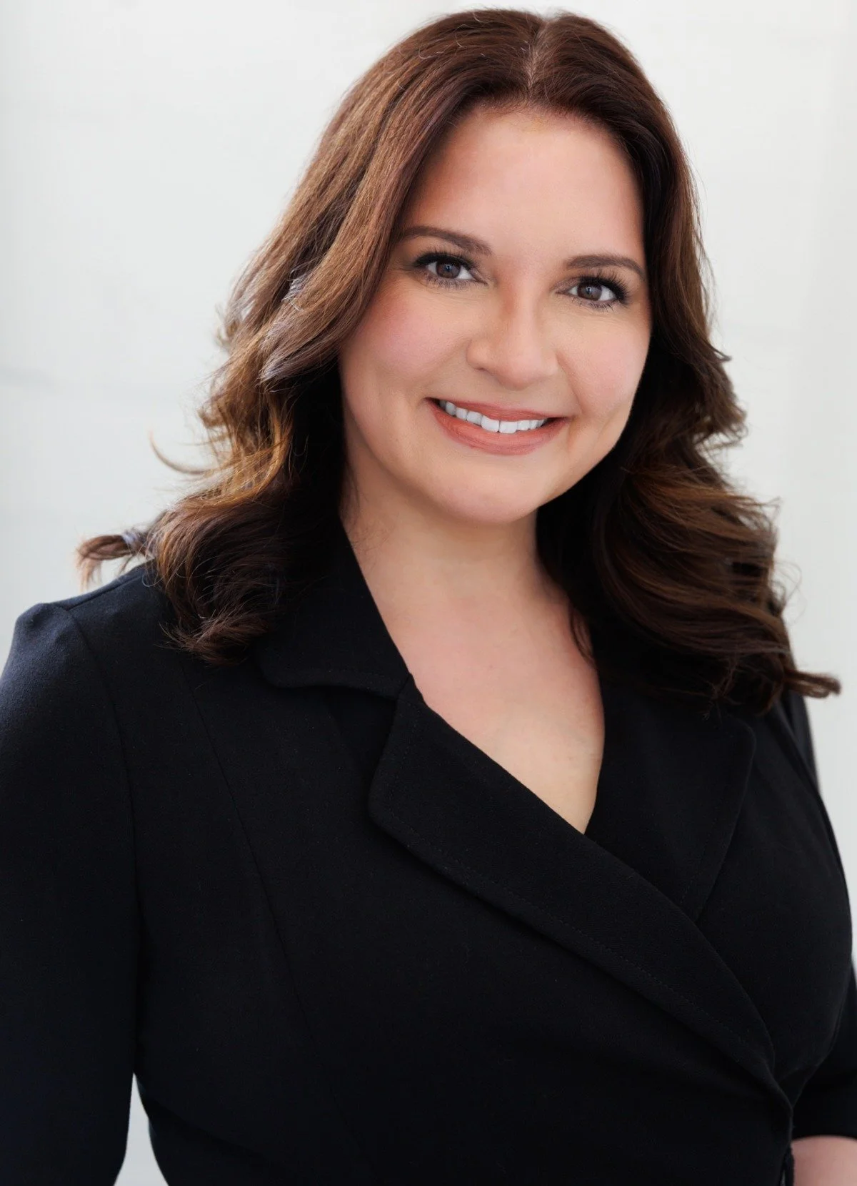 A woman with shoulder-length brown hair and light makeup smiling in a black blazer against a neutral background.
