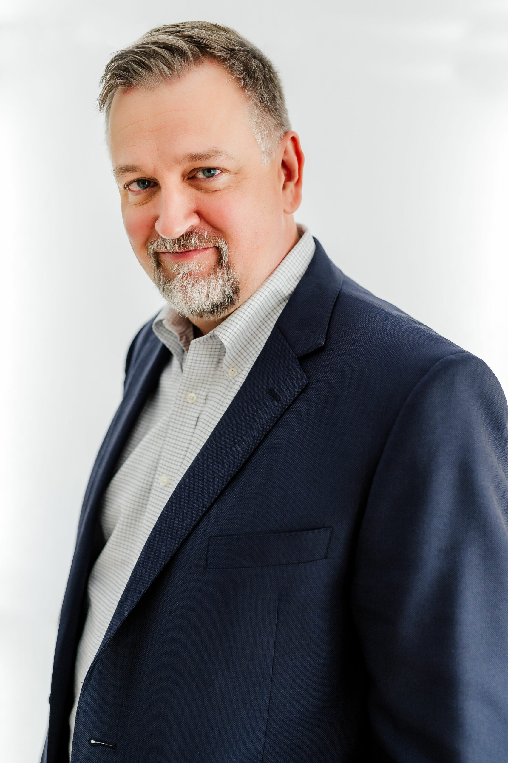A middle-aged man with gray hair, beard, and mustache wearing a dark blazer and a checkered shirt, standing against a plain white background.