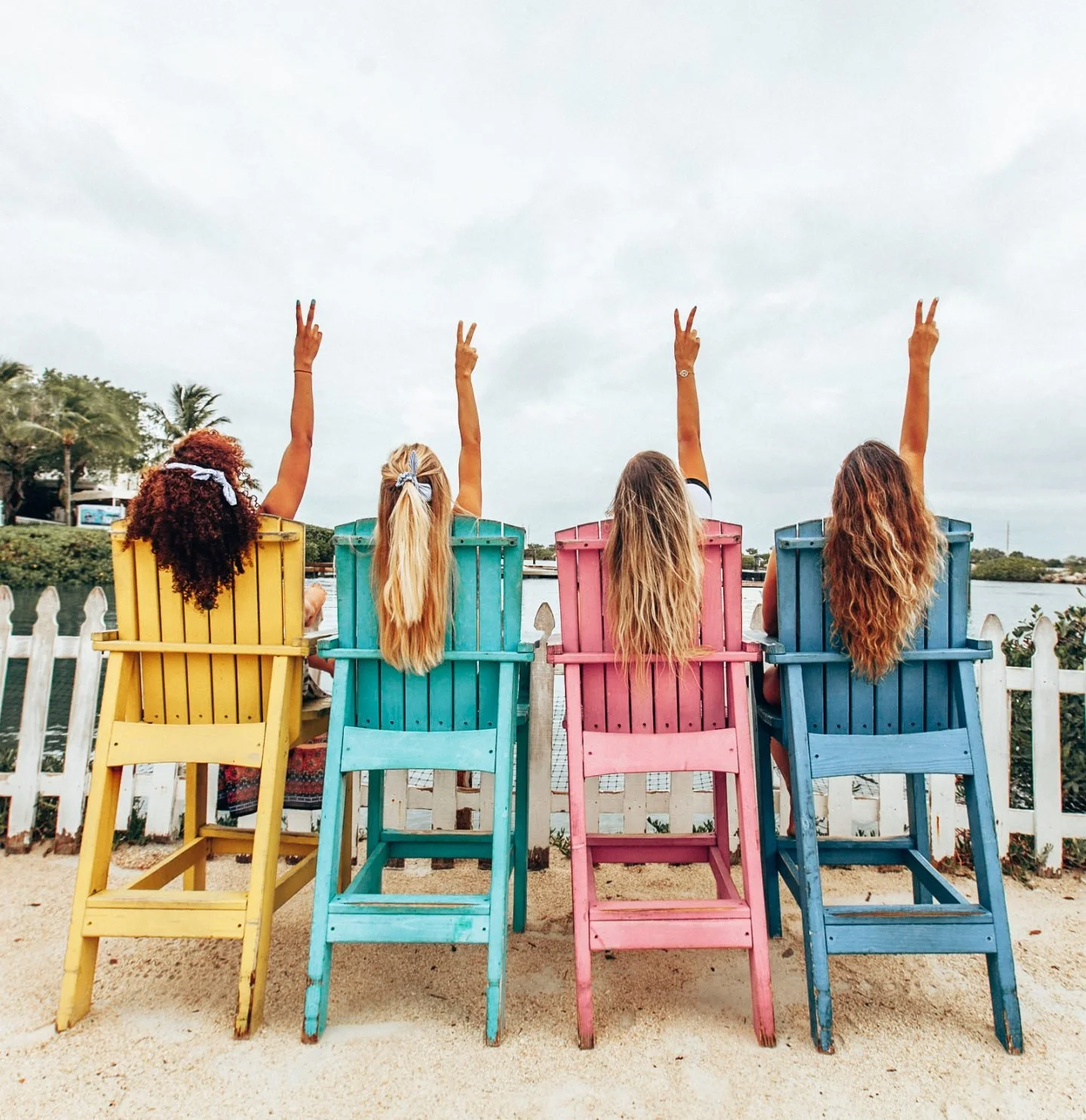 Four women sitting on colorful chairs by a fence, facing water, with their arms raised making peace signs, and enjoying a cloudy day.