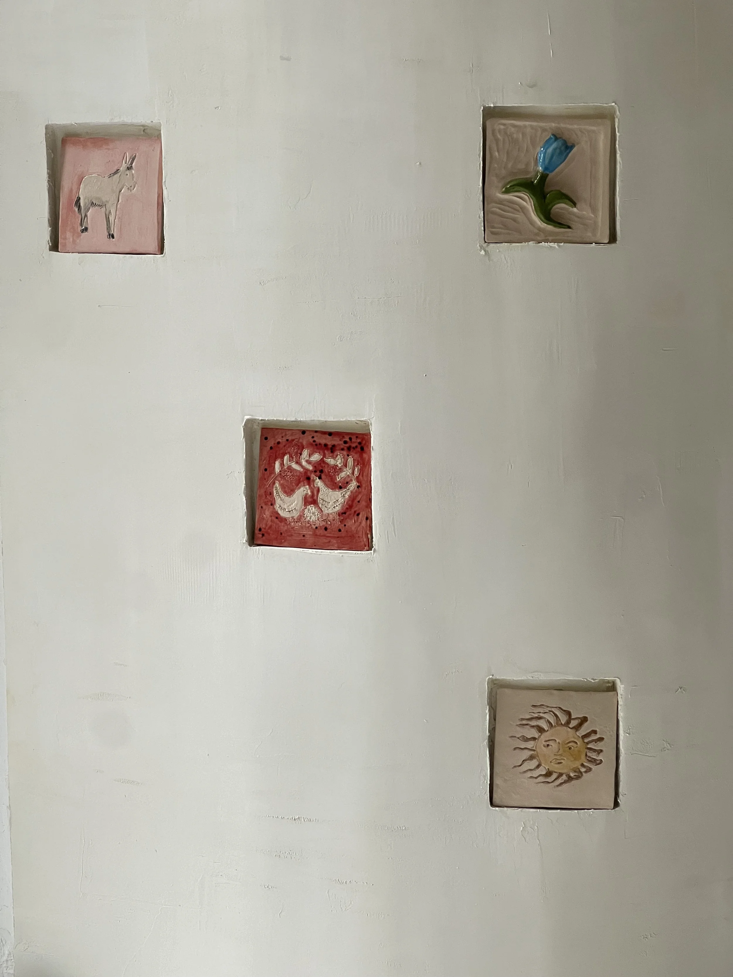 Quatre carreaux décoratifs encastrés dans un mur blanc, chacun avec un dessin différent d'un animal ou d'un symbole : un âne, une tulipe, deux colombes et un soleil avec un visage.
