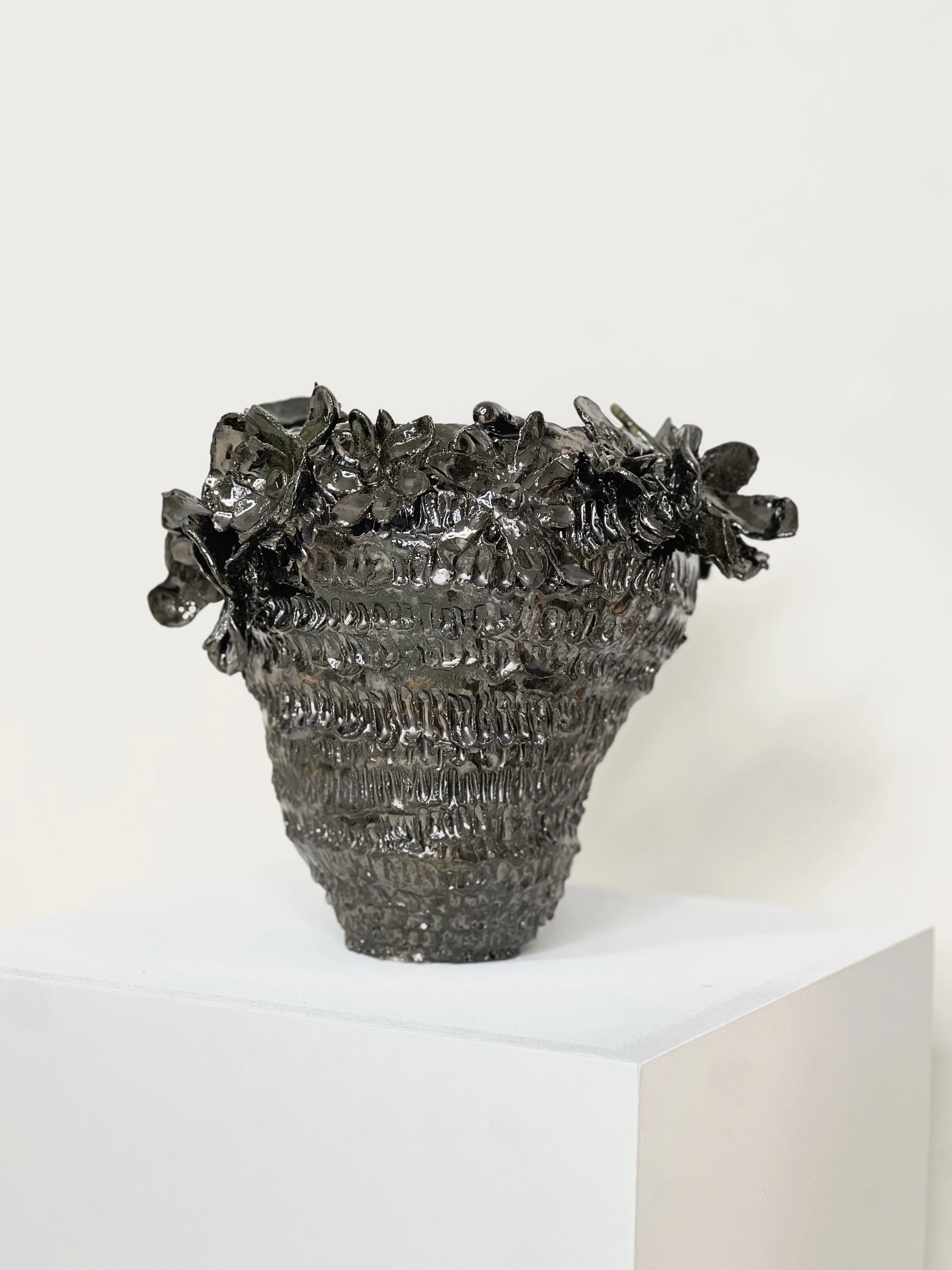 Vase en métal noir avec décor floral en haut, exposé sur un socle blanc